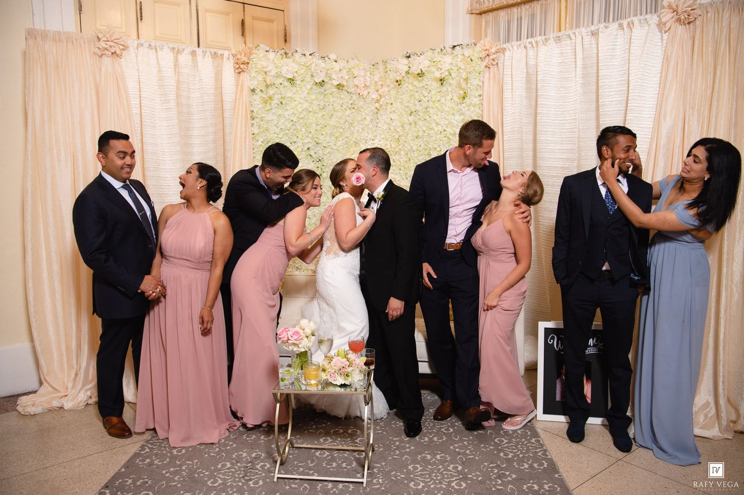 Wedding party celebrates with kisses at floral backdrop, bridesmaids in blush pink dresses flanking the newlyweds.
