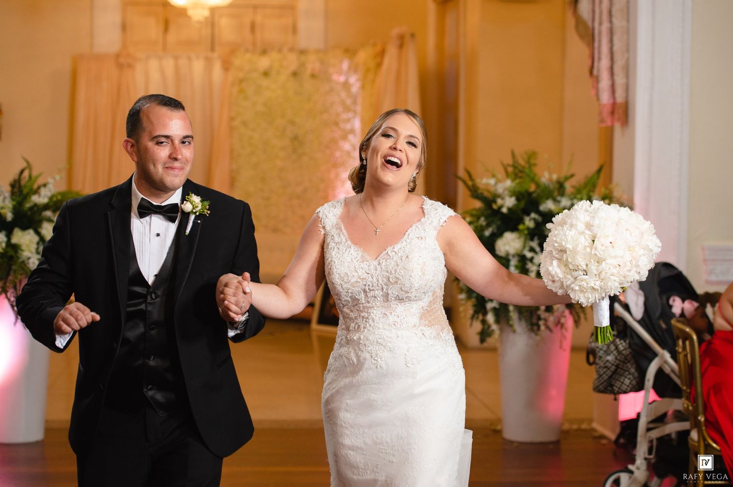 Joyful bride and groom holding hands walking into wedding reception, bride laughing holding white bouquet, groom in black tuxedo.