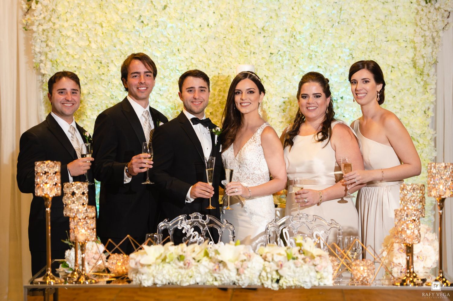 Wedding party group photo with bride, groom, and bridesmaids toasting champagne at floral reception.