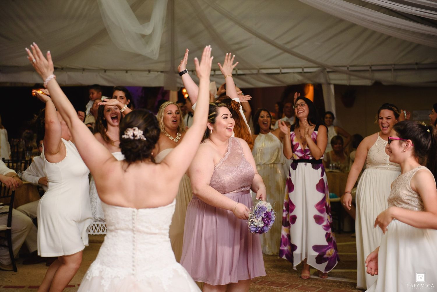 Bride tossing bouquet to excited guests at a wedding reception under a white tent.
