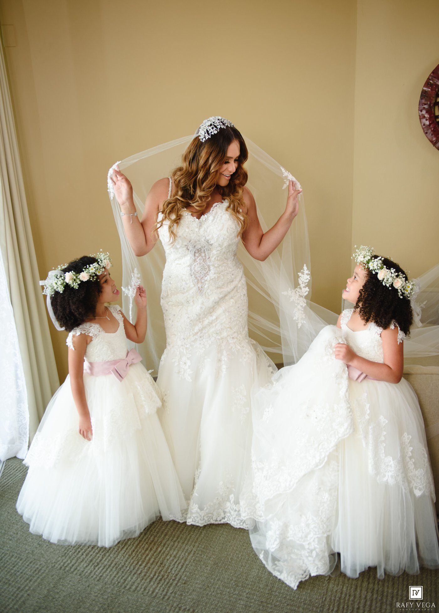 Bride in lace mermaid gown lifts veil while two flower girls in white dresses stand beside her.
