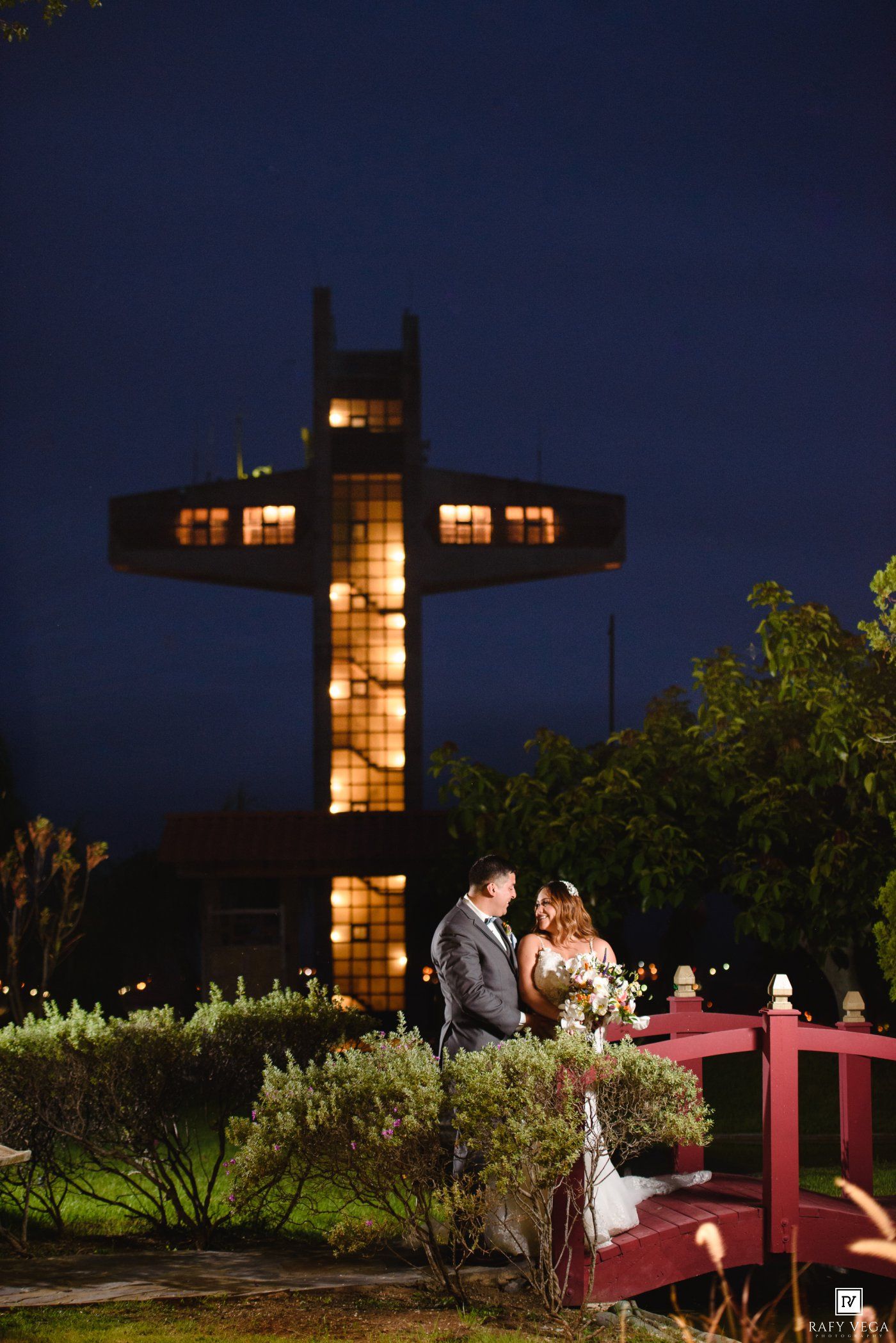 Bride and groom share a romantic moment at night with an illuminated cross monument glowing behind them.