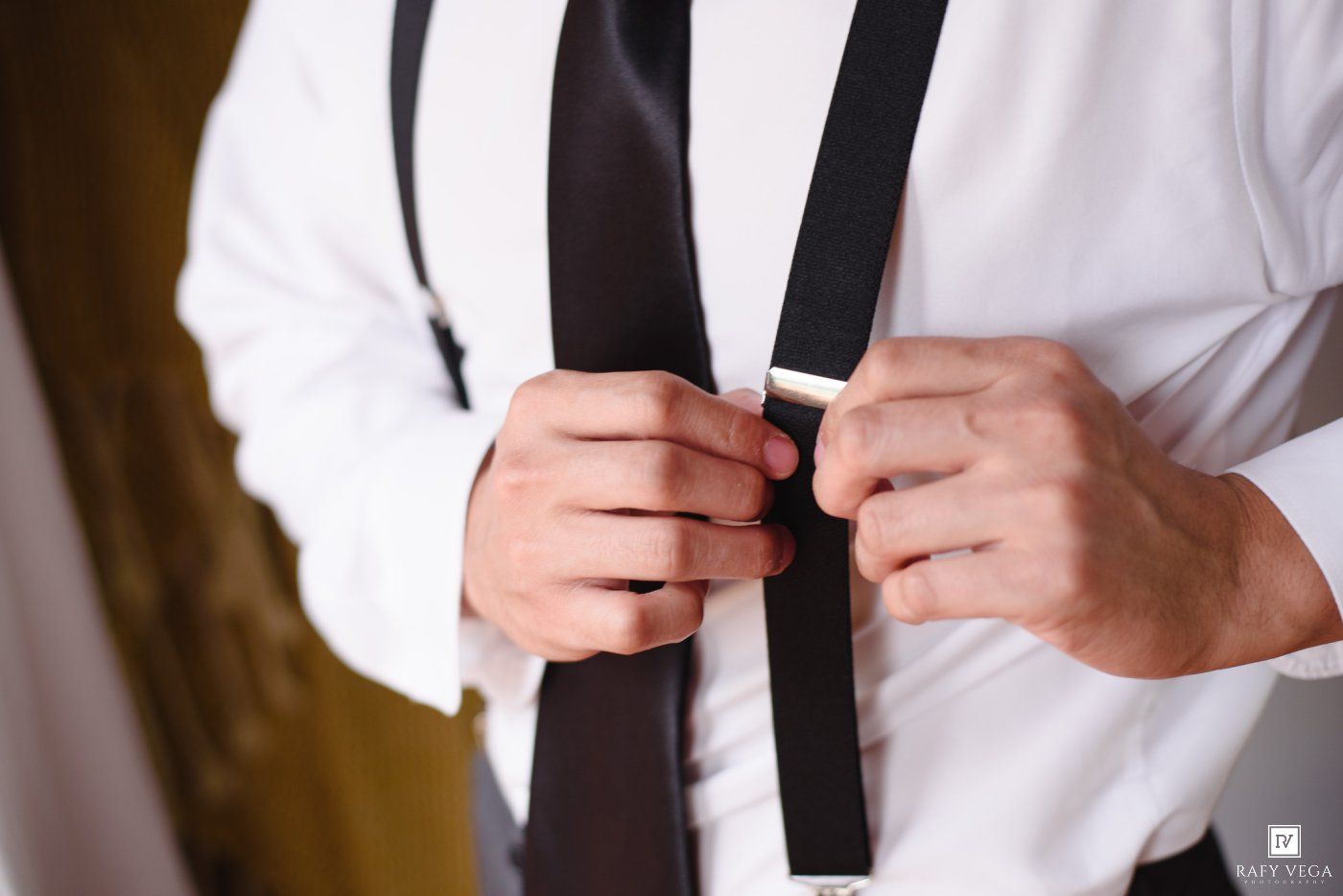 Man in white dress shirt adjusting black suspenders and tie, getting dressed for formal occasion.