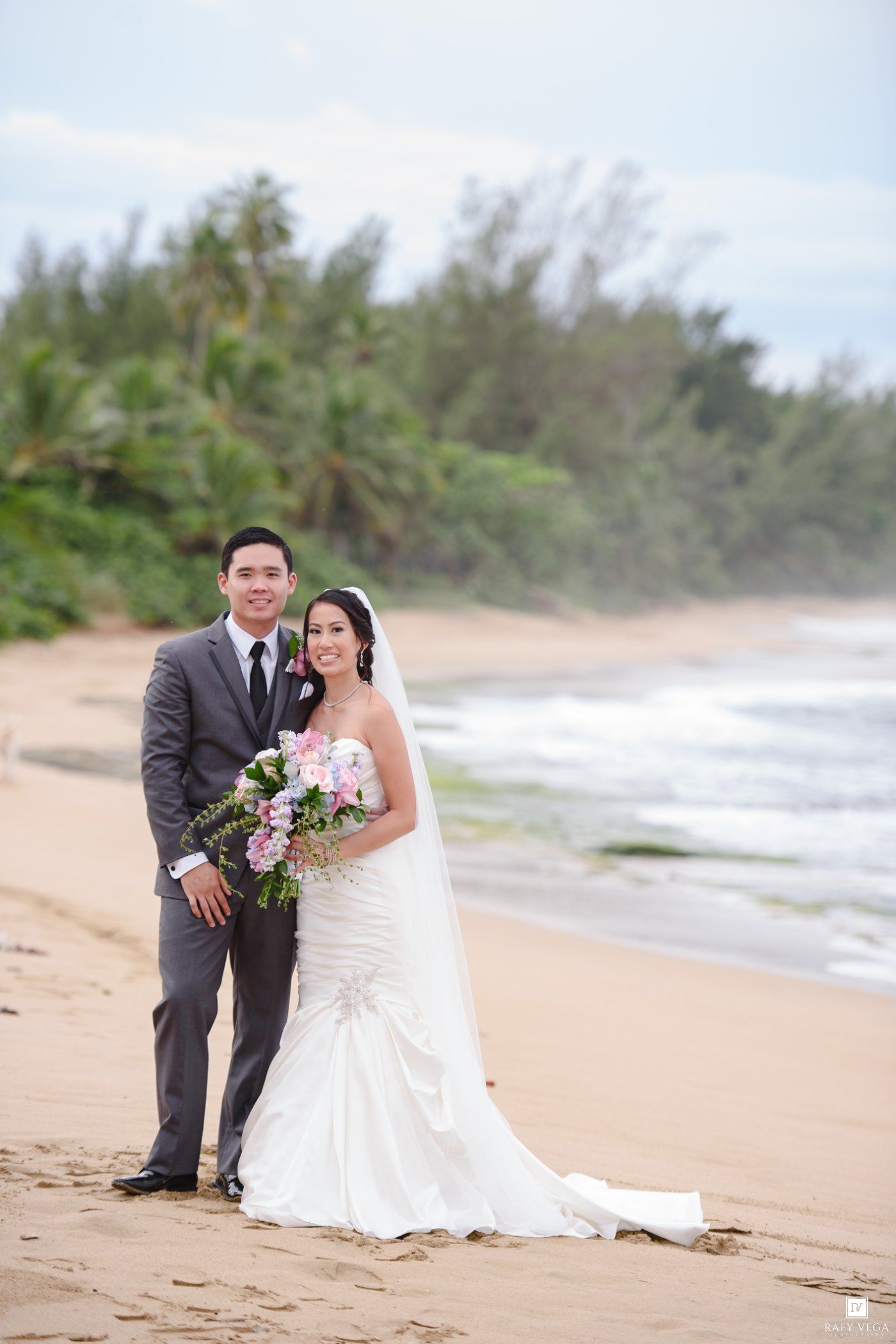 Bride and groom posing on a tropical beach, she holds a pink bouquet in a white gown, he wears a gray suit.