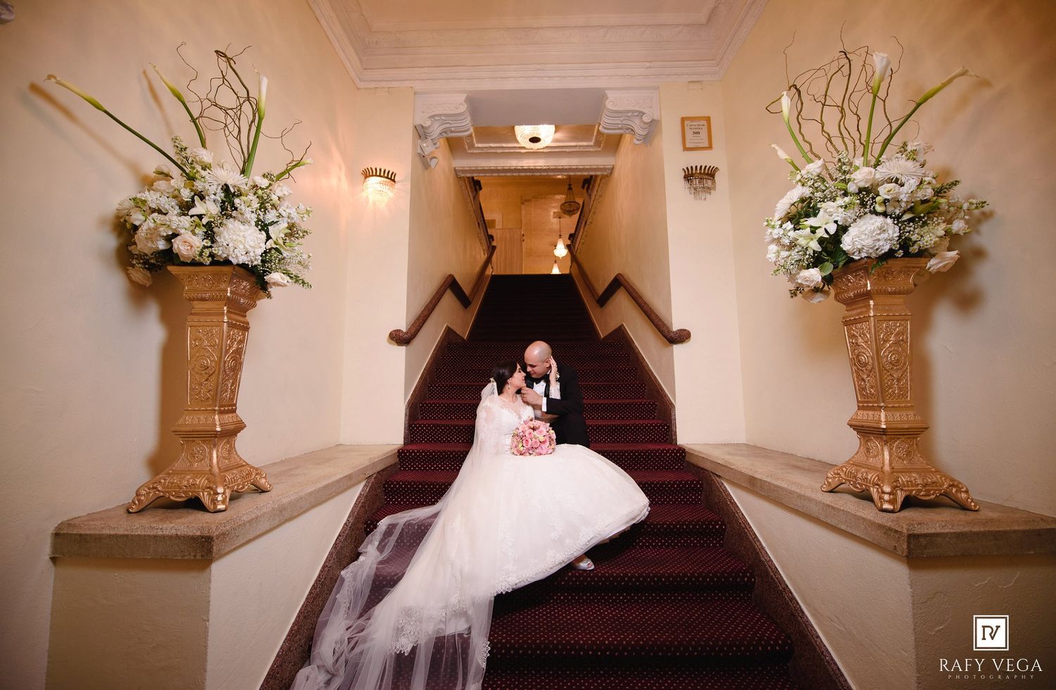 Bride in white gown sitting on red carpeted staircase flanked by tall floral arrangements in elegant venue.