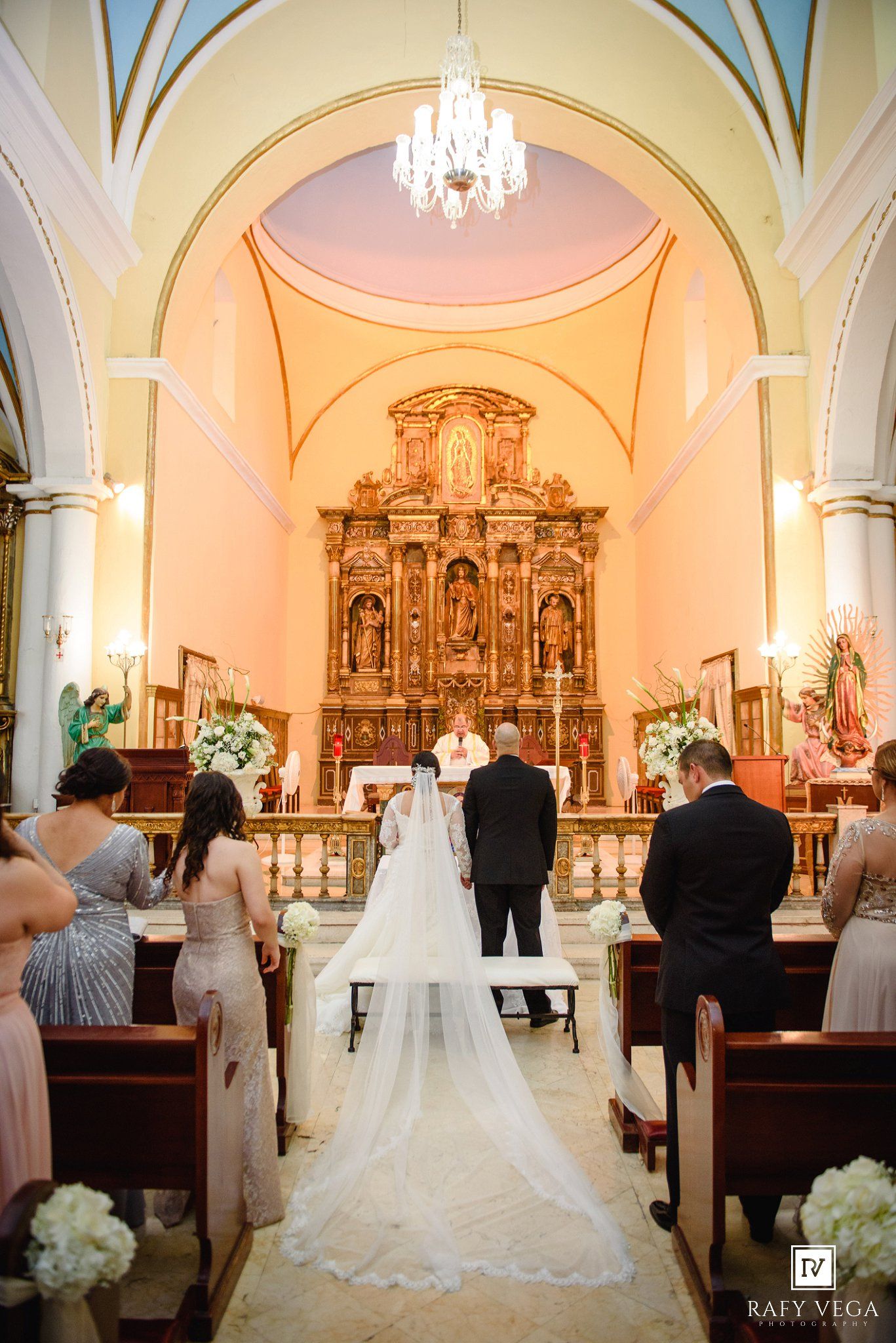 Wedding ceremony in ornate church with bride and groom at altar before golden altarpiece.