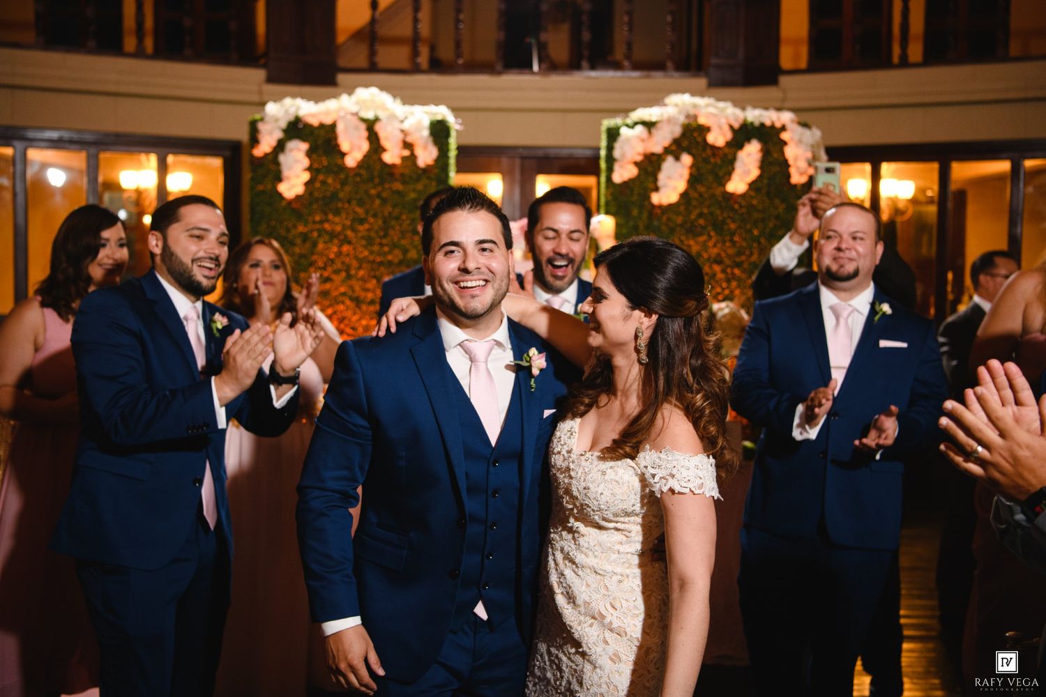 Smiling bride and groom celebrate at elegant wedding reception with groomsmen clapping in background near floral wall.