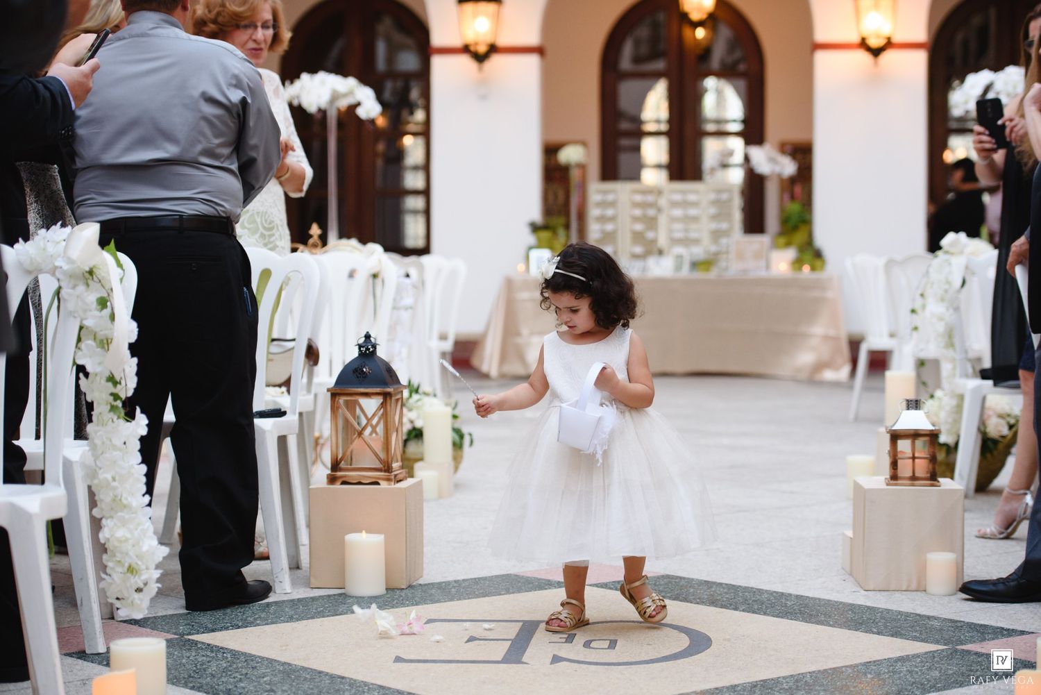 A flower girl in a white dress walks down the aisle at an elegant outdoor wedding ceremony with candles and white florals.