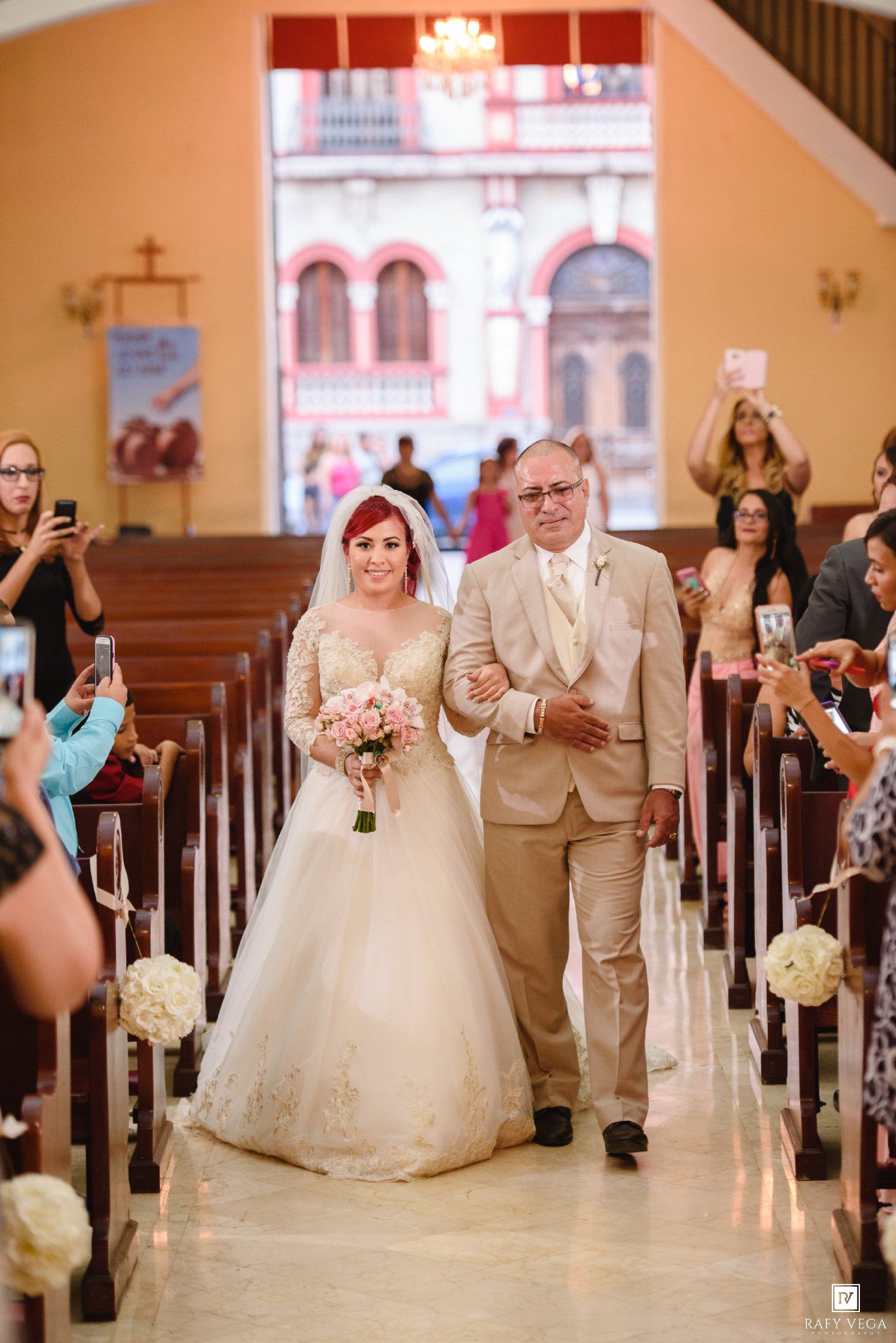 Bride in white gown walks down church aisle with older man in tan suit, guests watching and photographing.