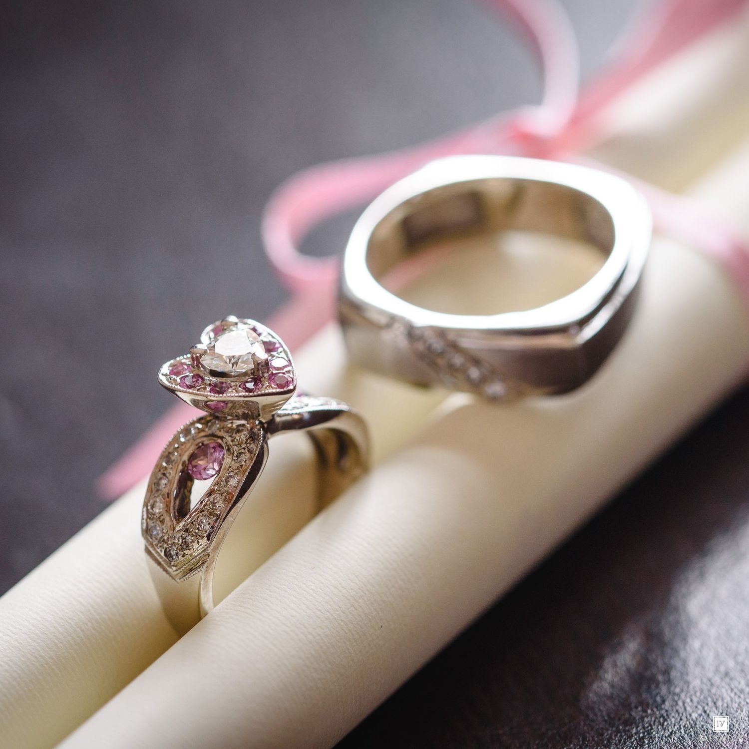 Two elegant silver rings with pink and white gemstones displayed on a cream roll with a pink ribbon background.