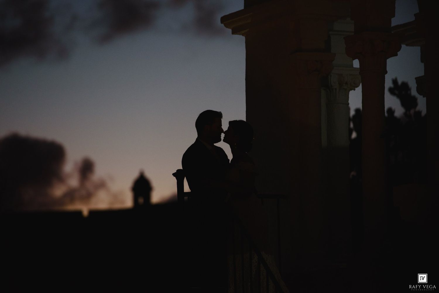 Silhouette of a romantic couple kissing at sunset near historic architecture with a colorful dusk sky.