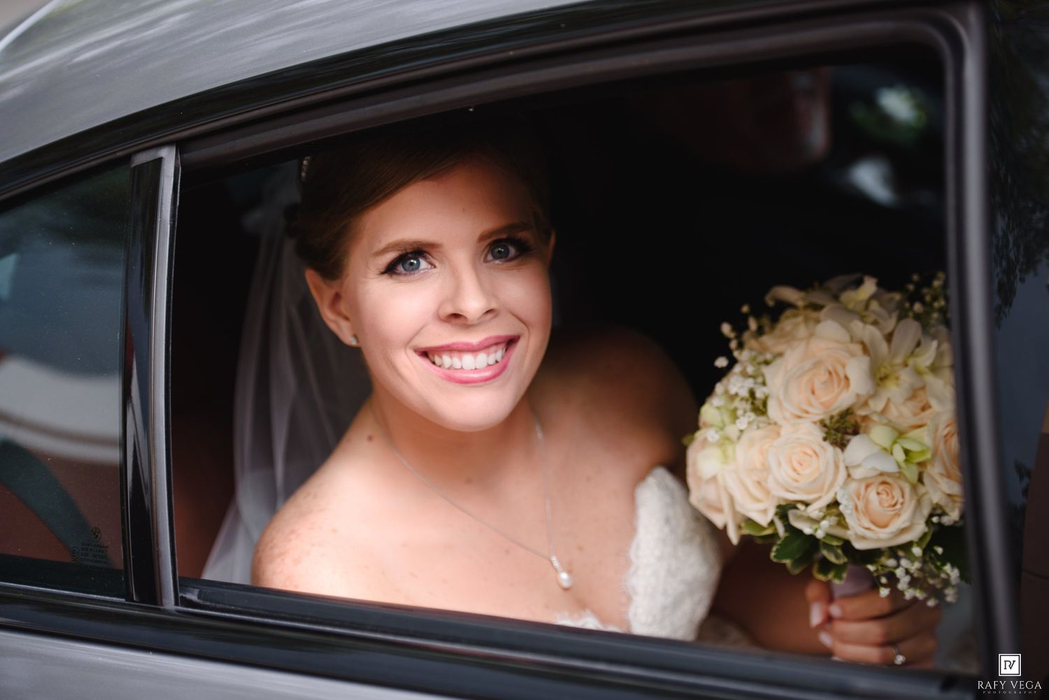 Radiant bride smiling in car window holding elegant white rose bouquet on her wedding day.
