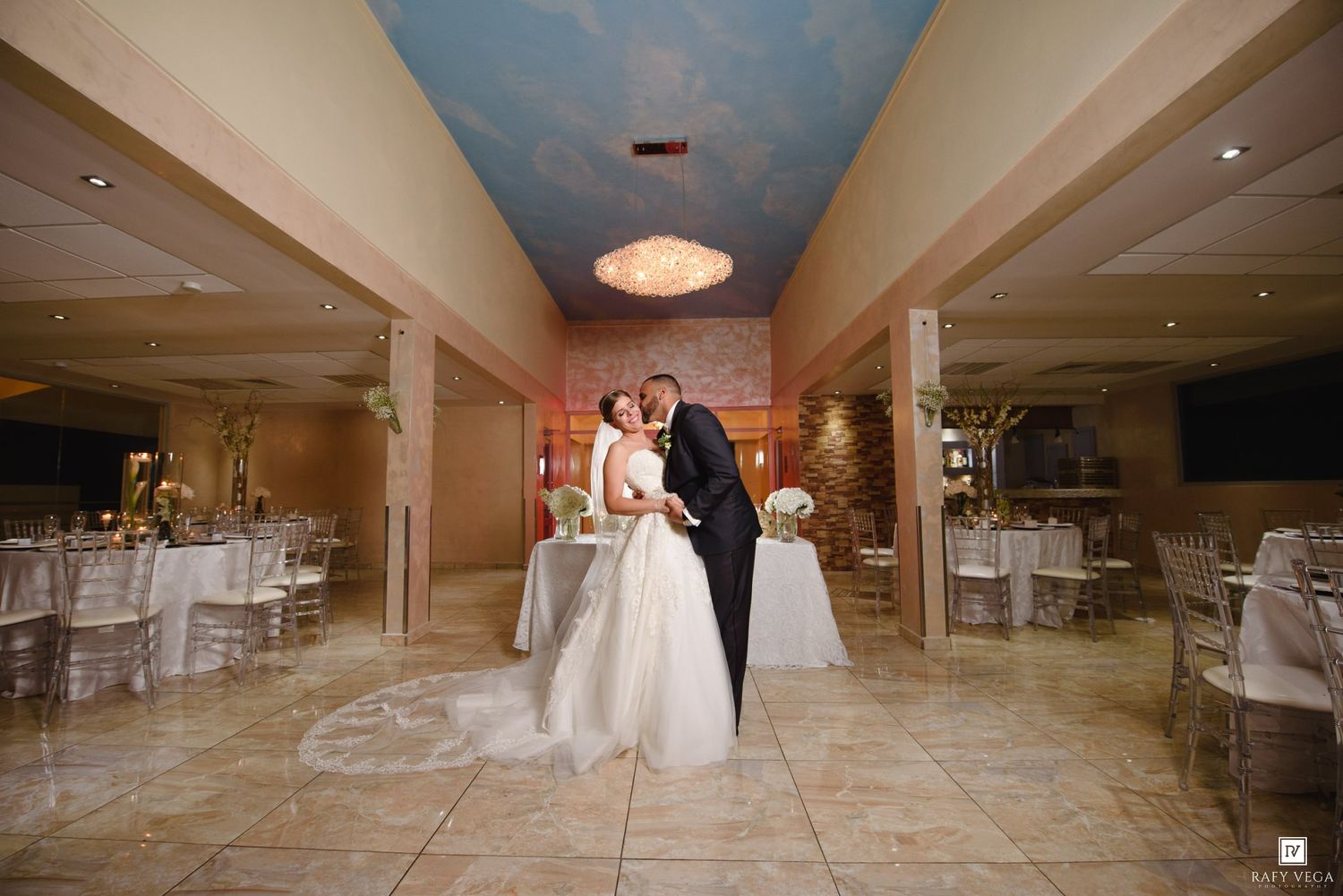 A bride and groom share a romantic kiss in an elegantly decorated wedding reception hall with blue ceiling.