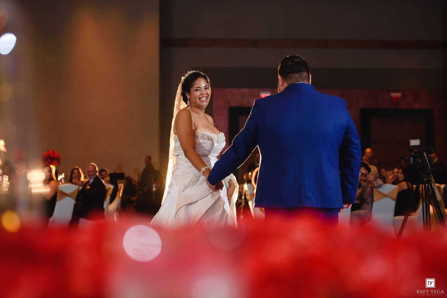Bride and groom share their first dance at a wedding reception with red floral arrangements and guests in the background.