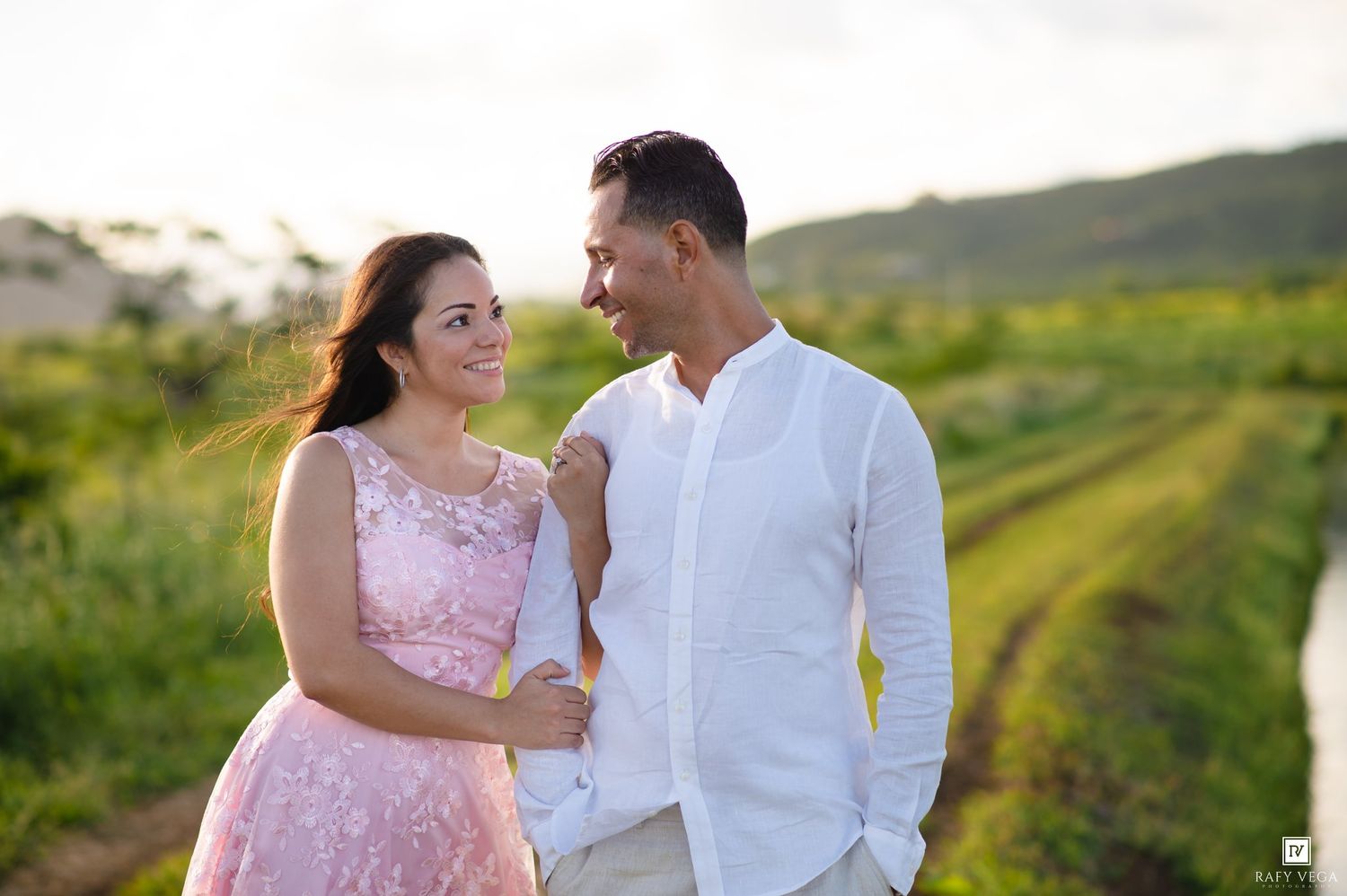Couple in love smiling at each other outdoors, woman in pink dress and man in white shirt, lush green field background.