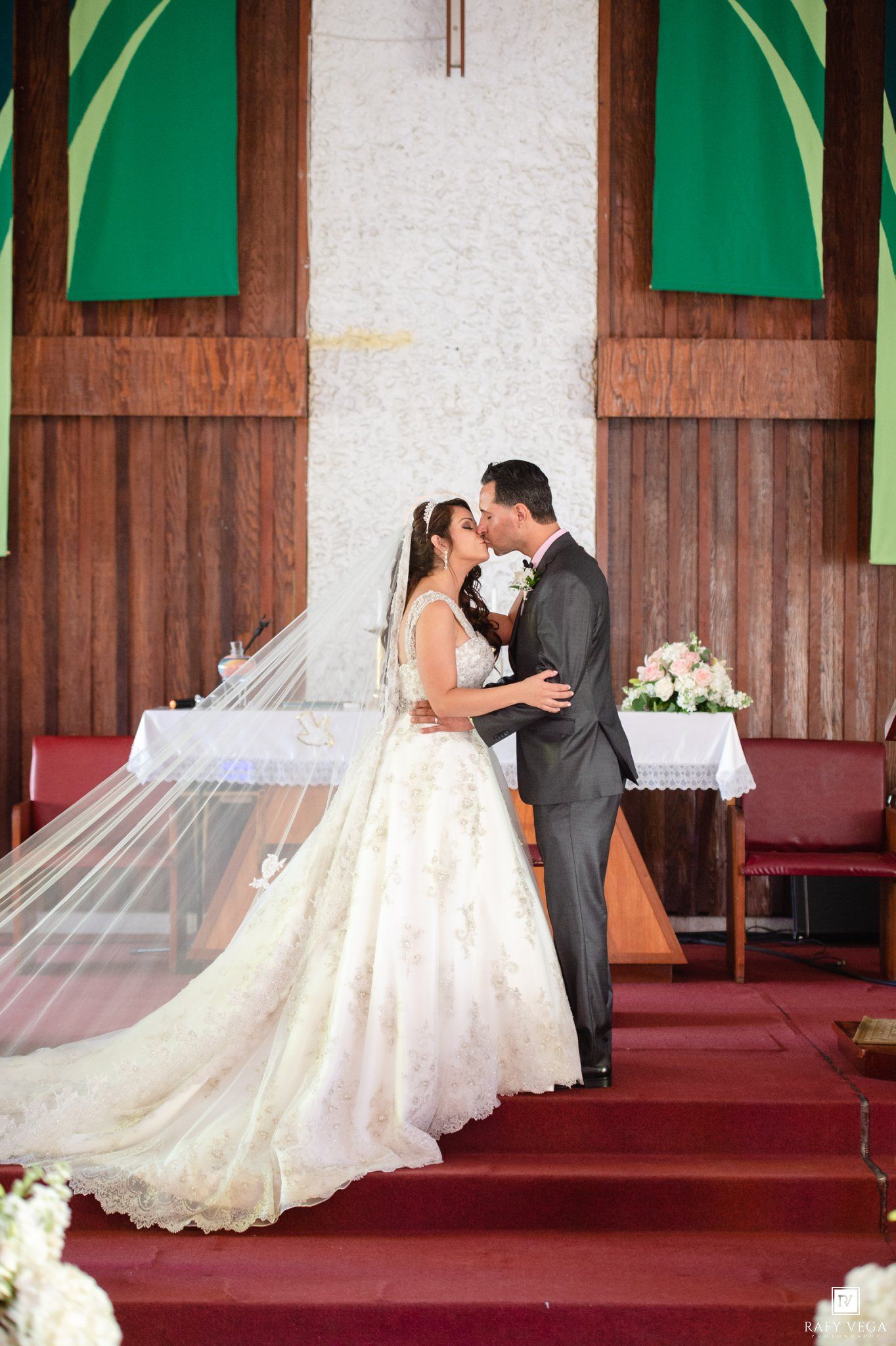 Bride and groom share first kiss at church altar, bride in lace gown with flowing veil, groom in gray suit.
