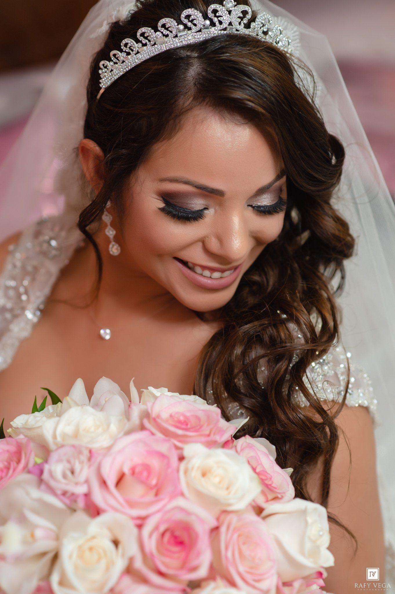 Bride wearing diamond tiara and veil holds pink rose bouquet, showcasing elegant bridal makeup and curled hair.