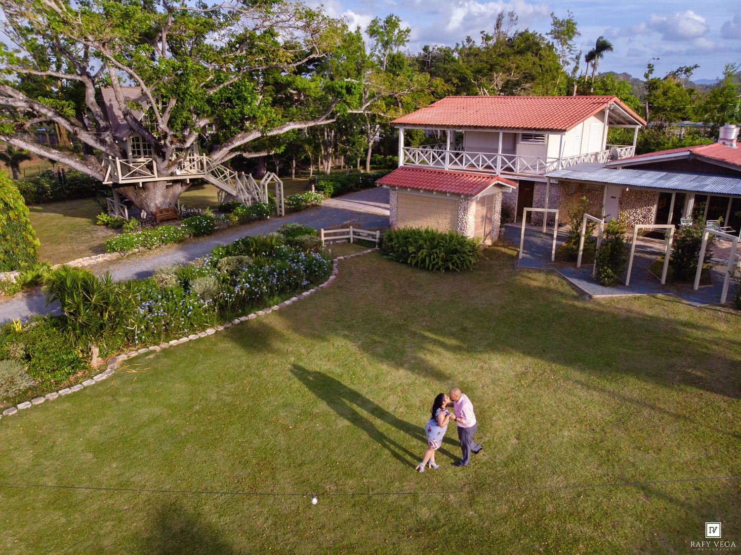 Aerial view of a couple embracing on a lush green lawn beside a charming red-roofed tropical estate.