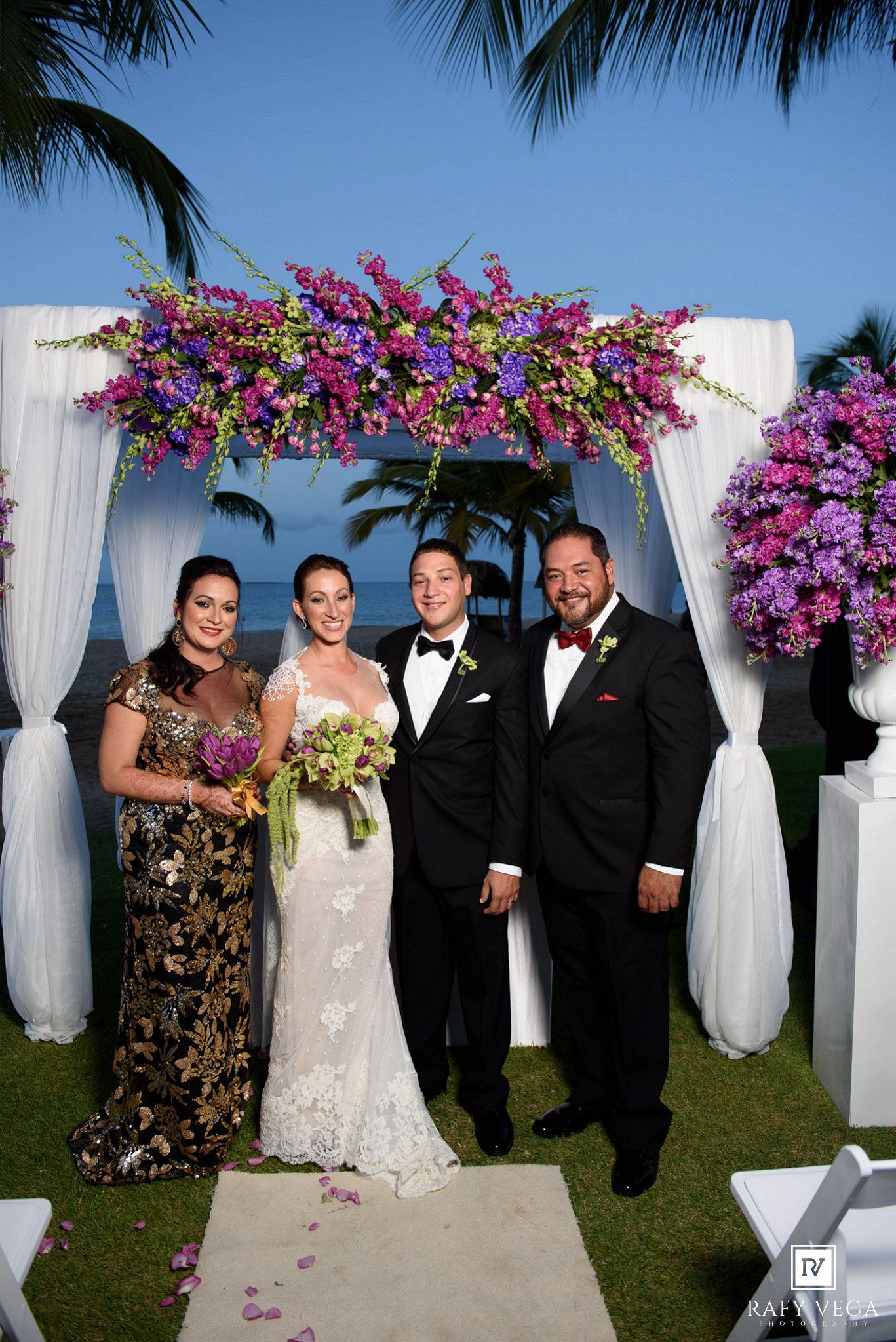 Bride and groom pose with guests under a purple floral arch at an outdoor beach wedding ceremony.