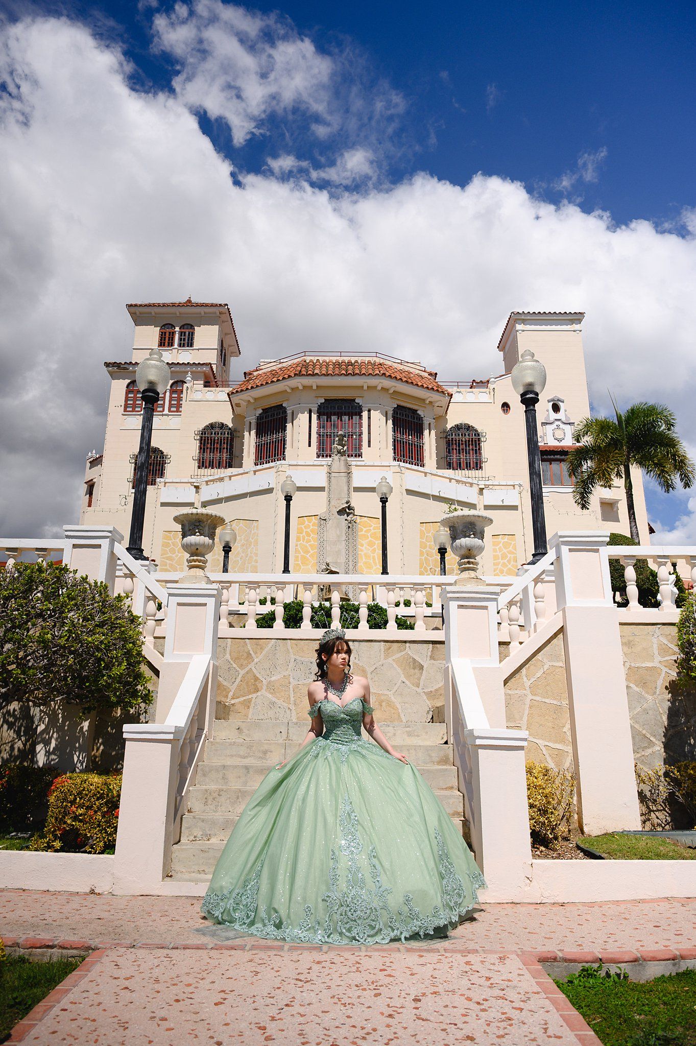 Young woman in mint green quinceañera ballgown poses on white staircase before elegant Spanish-style mansion.