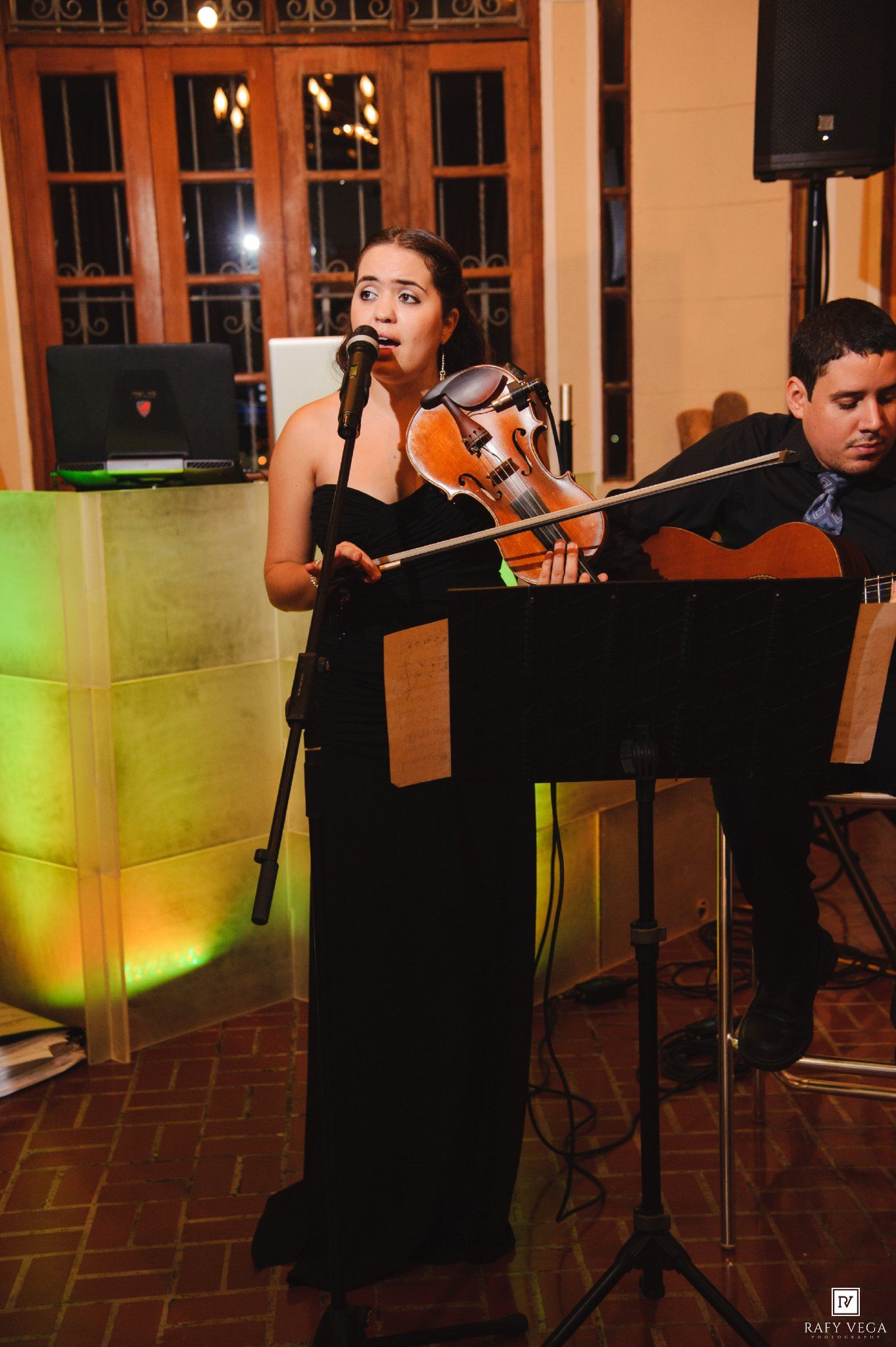 A woman in a black gown sings into a microphone while holding a violin, with a guitarist performing beside her.