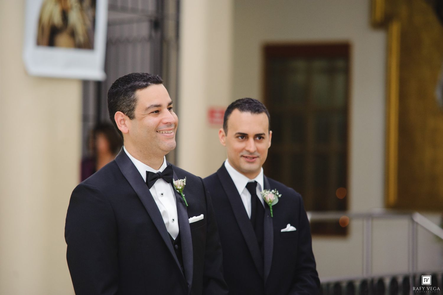 Two grooms in black tuxedos with white boutonnieres smile during their wedding ceremony inside a church.