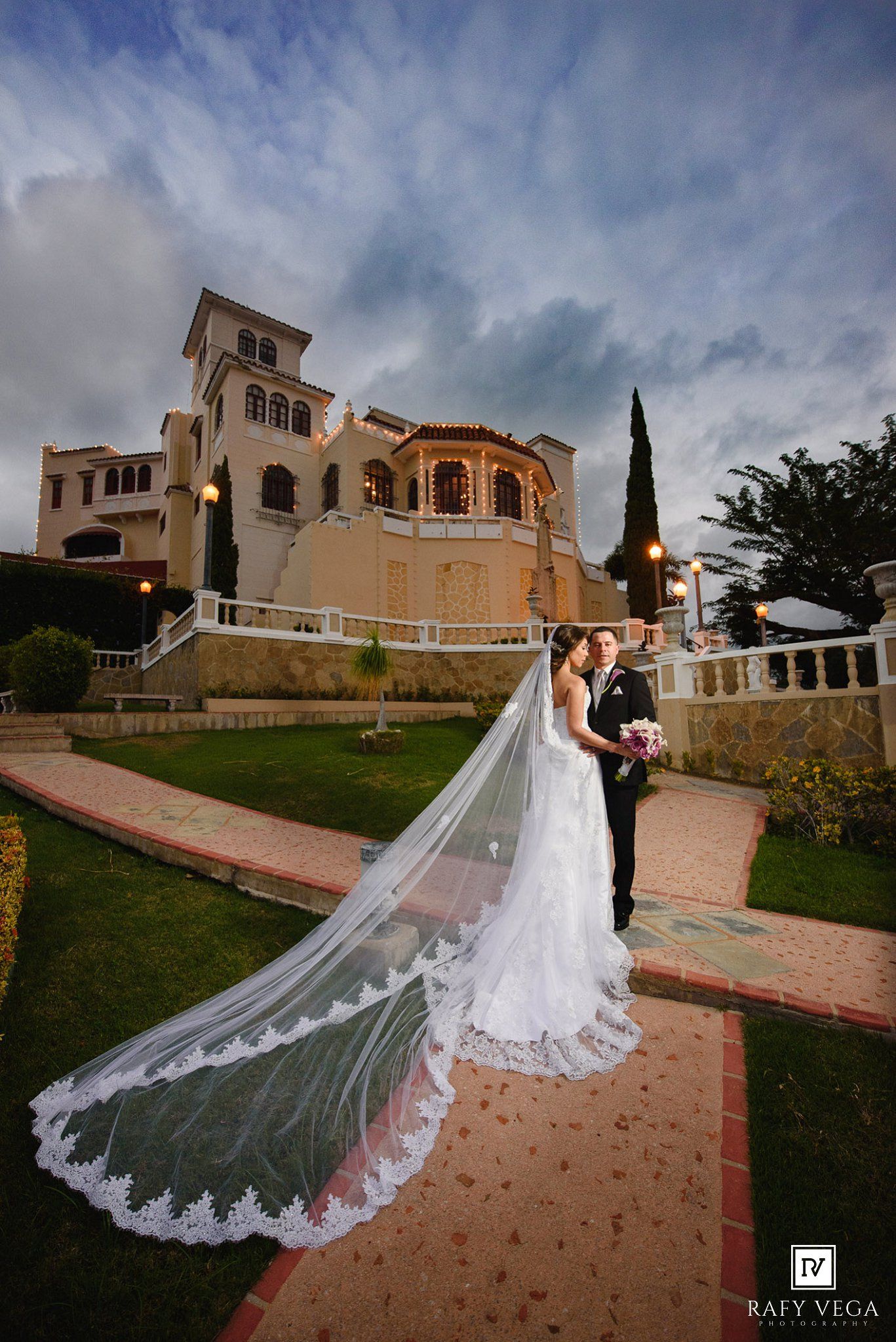 Bride and groom pose in garden of elegant Spanish villa with dramatic cloudy sky and long lace cathedral veil.