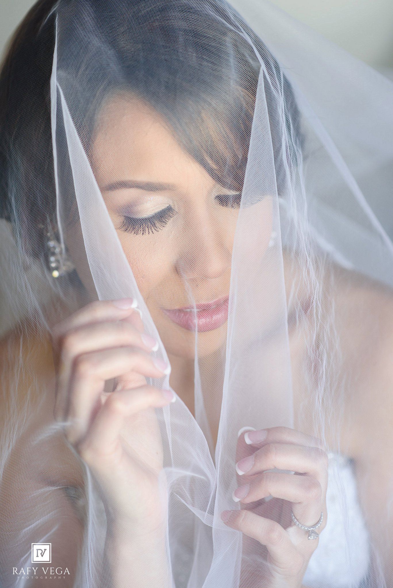 Bride in white veil looking down, showcasing elegant wedding makeup with long lashes and soft pink lips.