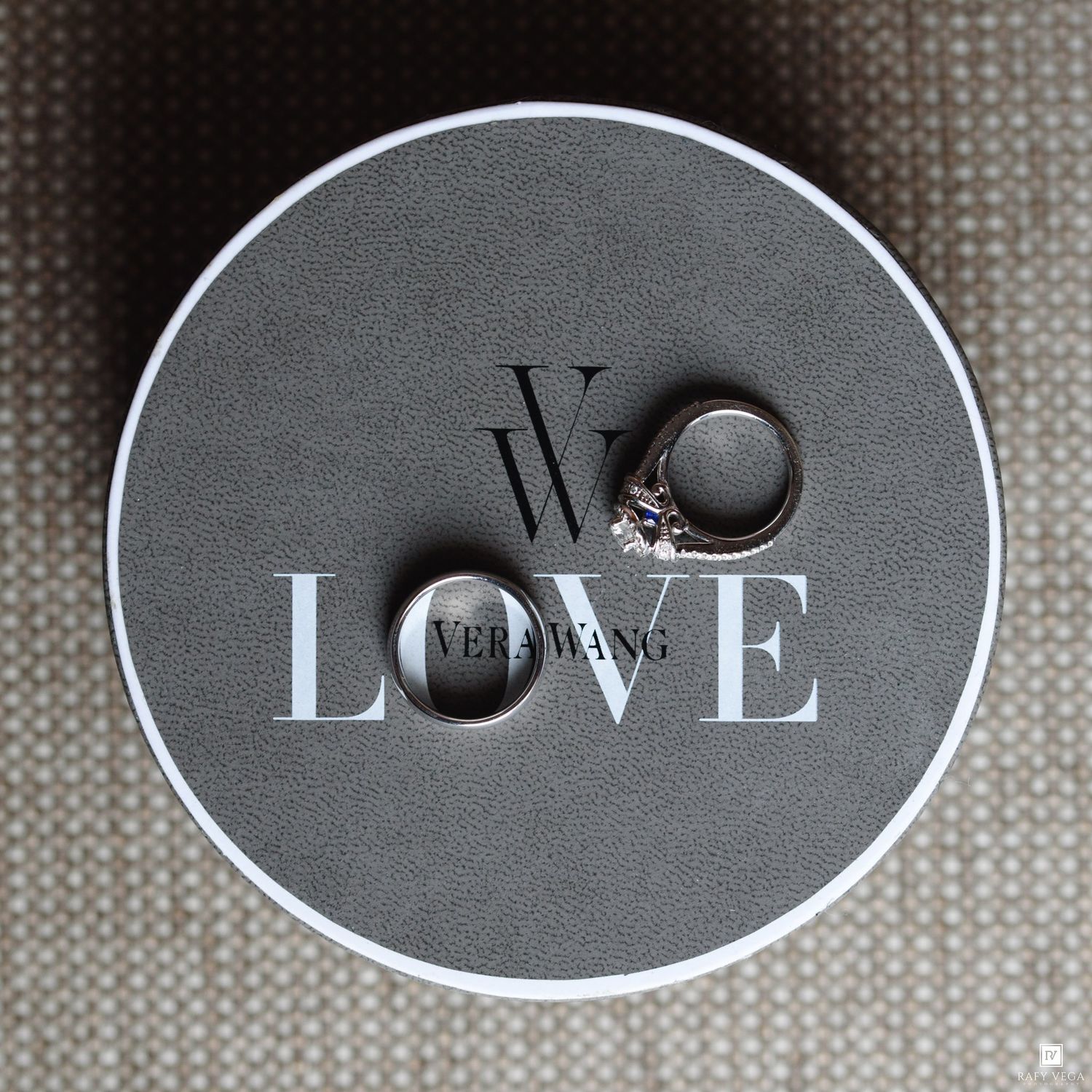 Two wedding rings displayed on a round Vera Wang Love jewelry box, featuring a diamond engagement ring and a plain band.