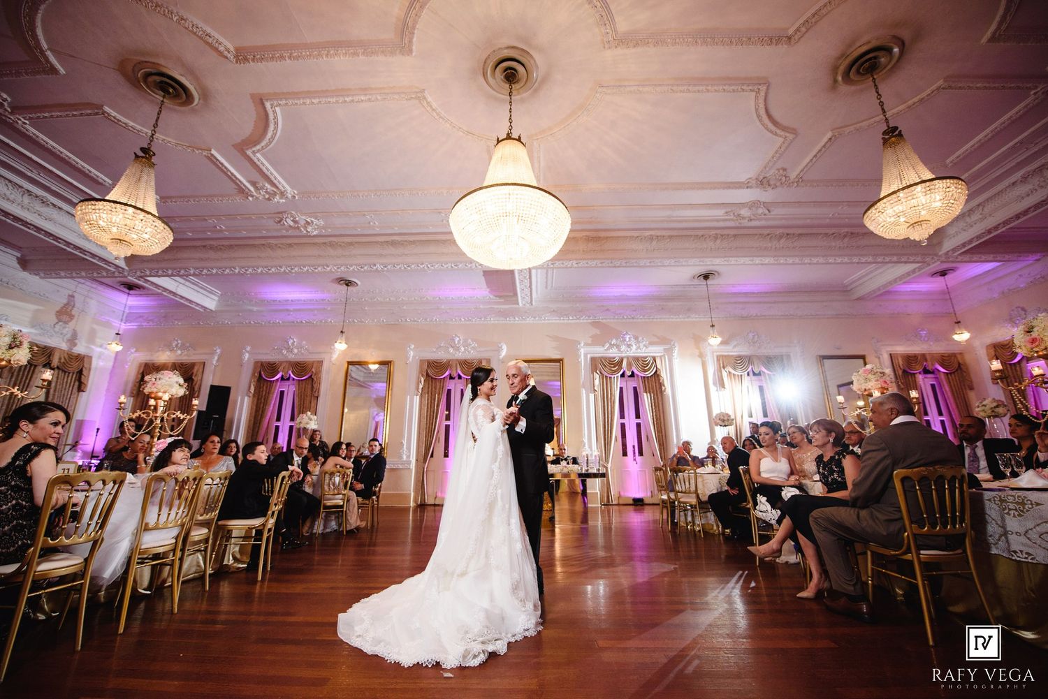 Bride and groom share first dance in ornate ballroom with gold chairs and purple uplighting at wedding.