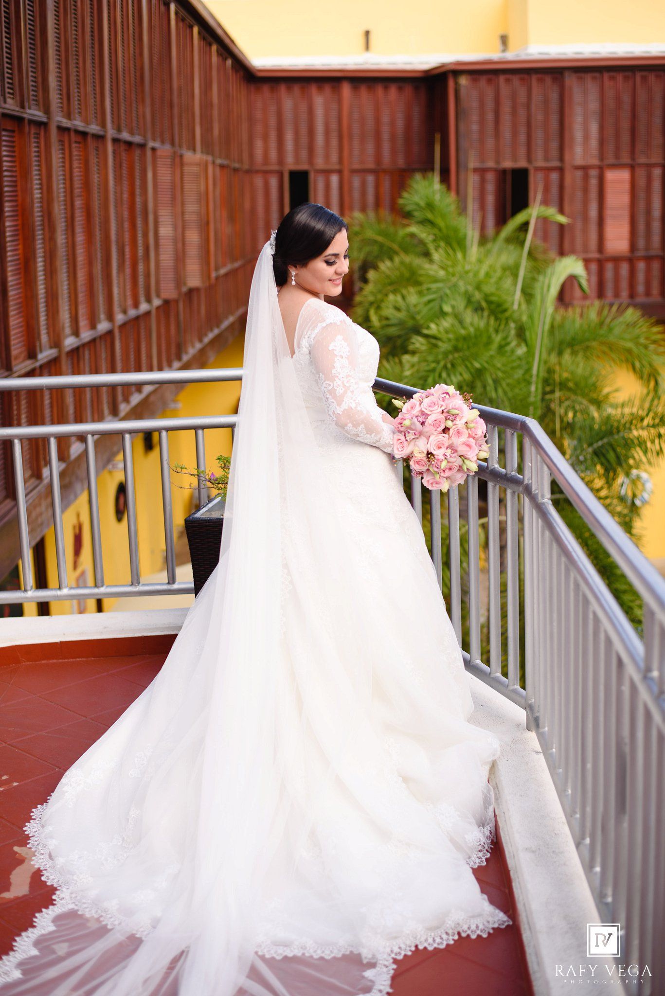 Bride in elegant long-sleeve white gown with veil and pink bouquet poses on balcony railing at wedding venue.