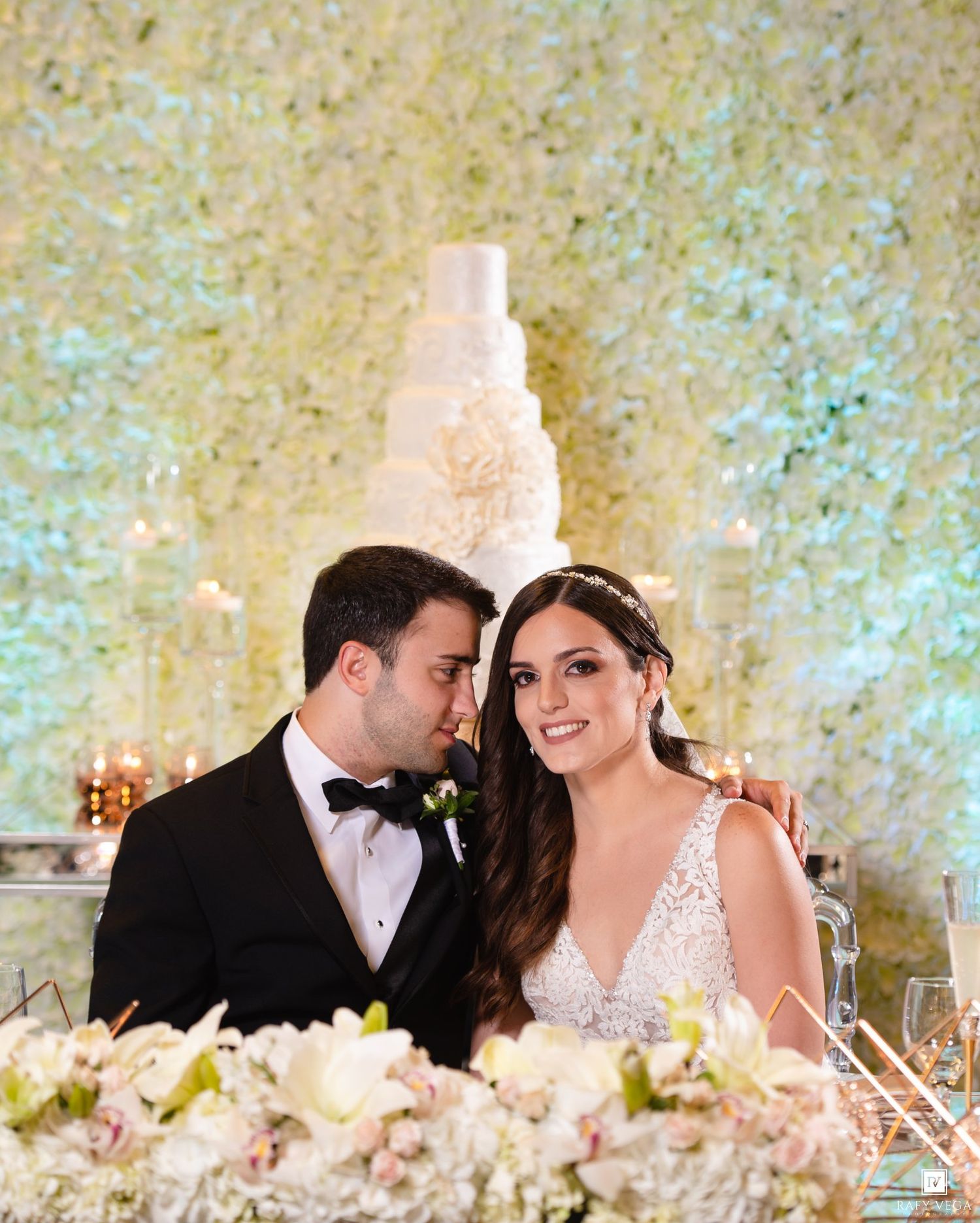 Couple in wedding attire smiling at sweetheart table with white floral wall and tiered cake backdrop.