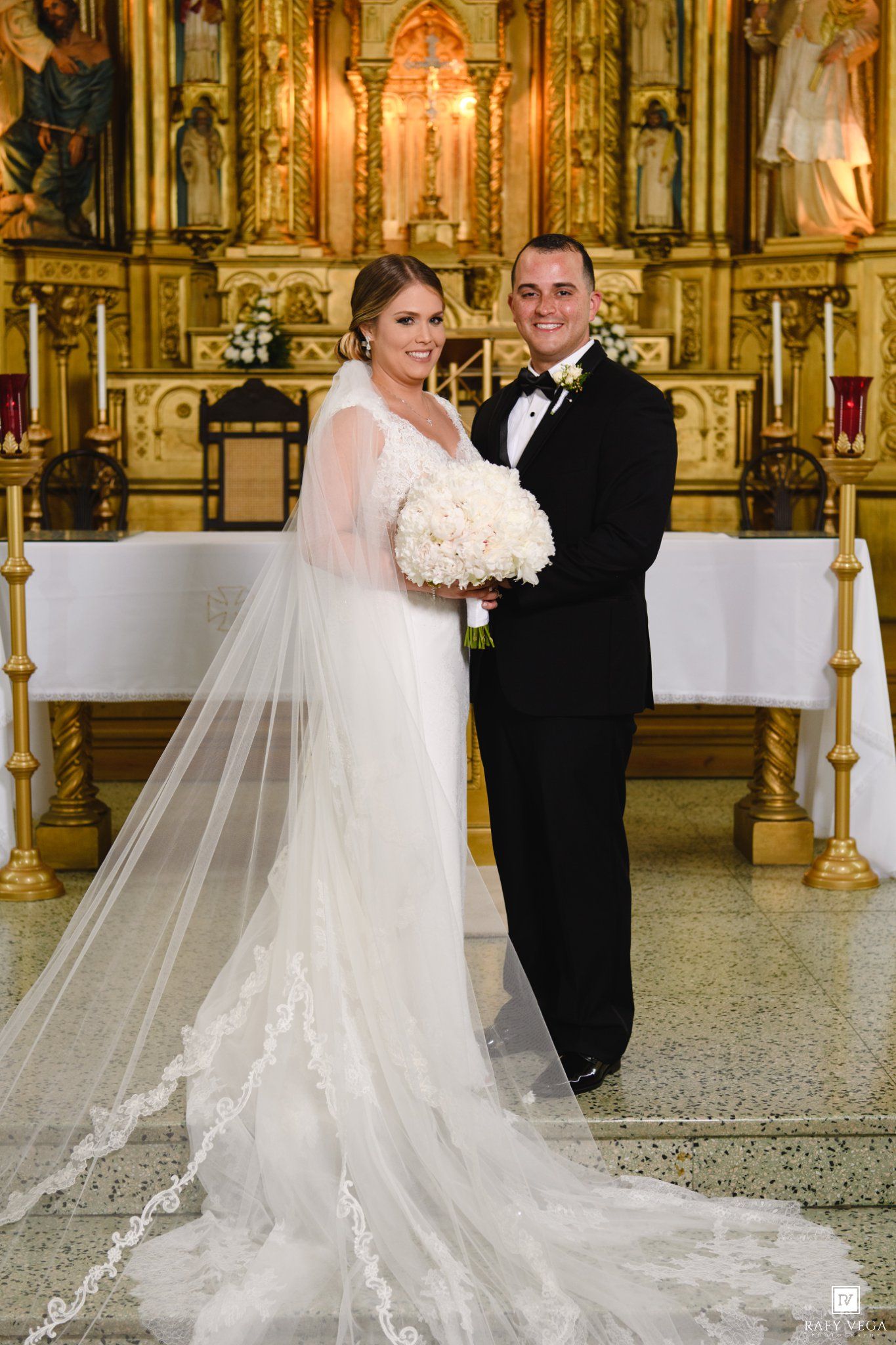 Newlyweds posing together in front of stunning golden church altar, bride holding white bouquet.