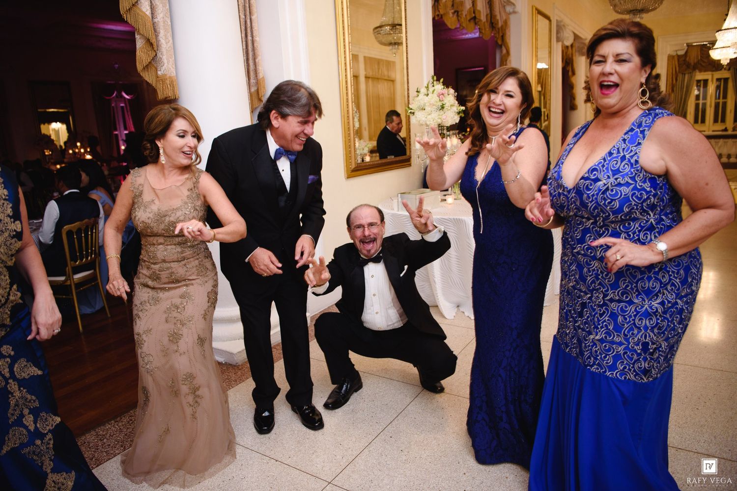 Guests celebrate joyfully at an elegant wedding reception, with one man doing a squat pose while others cheer.