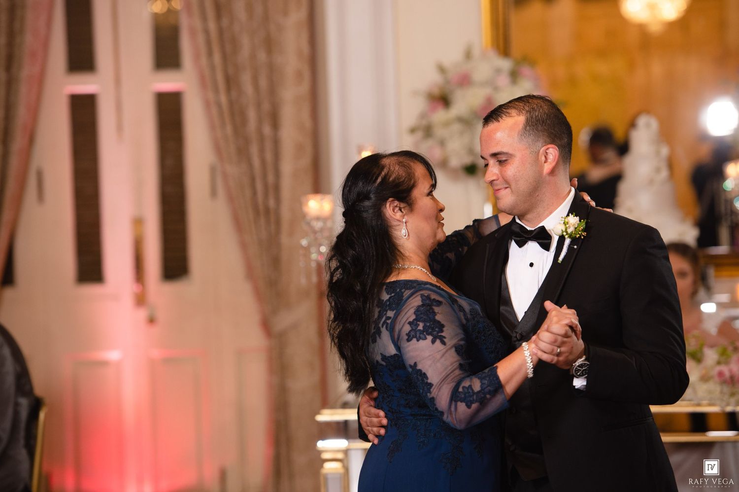Groom in black tuxedo shares mother-son dance with woman in blue lace gown at elegant wedding reception.