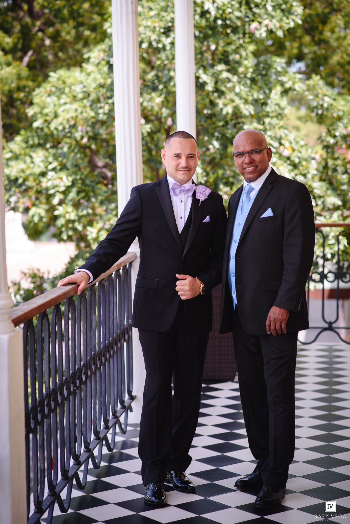 Two men in formal suits pose on a checkered balcony with white columns and lush green trees at a wedding.