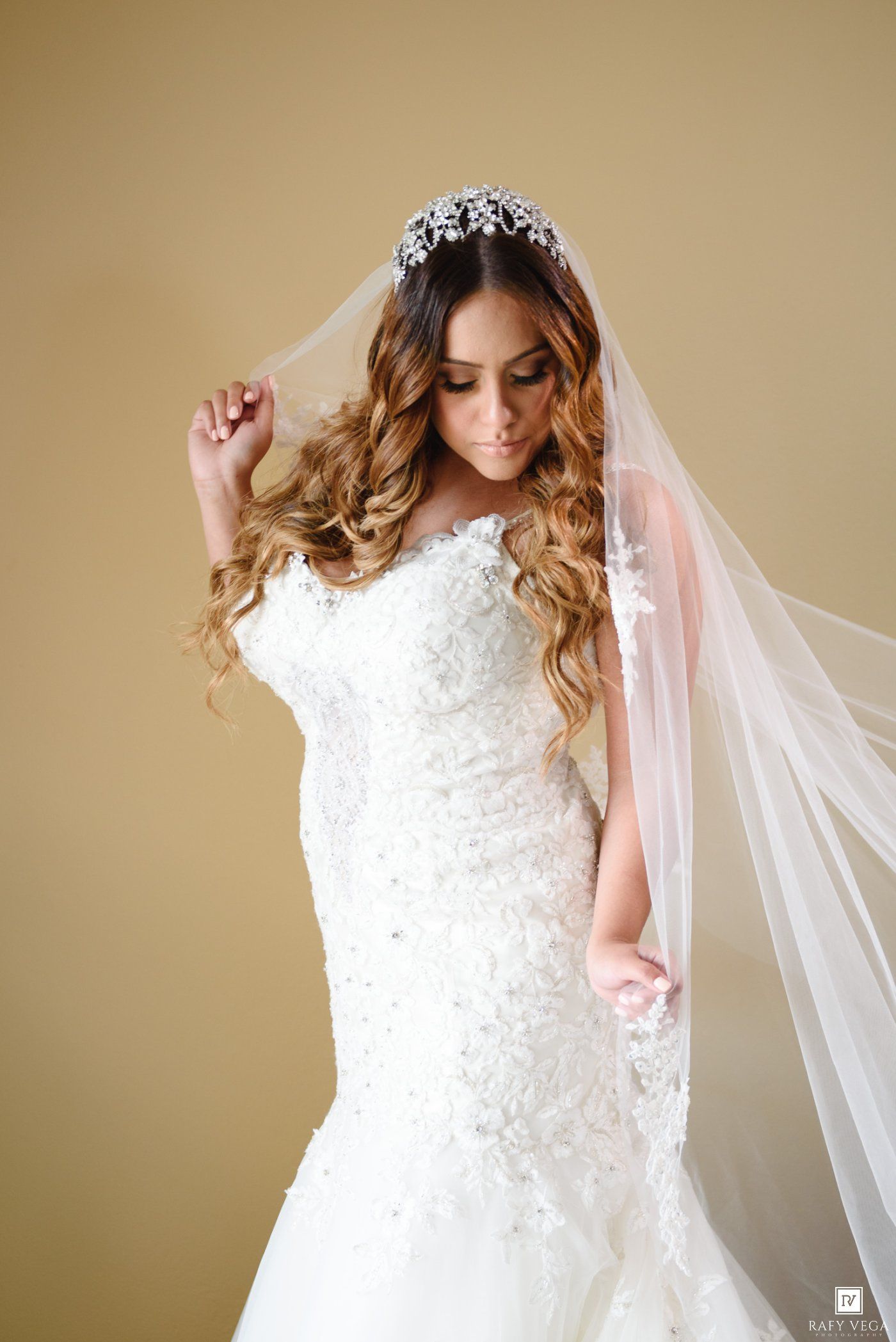 Bride in a lace mermaid wedding dress with a crystal tiara and flowing veil, posing elegantly against a gold background.