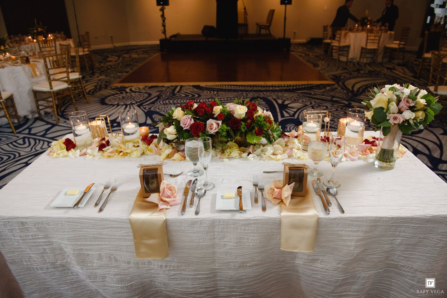 Elegant wedding reception head table with white linen, gold runners, floral centerpieces, and candlelight in a ballroom.