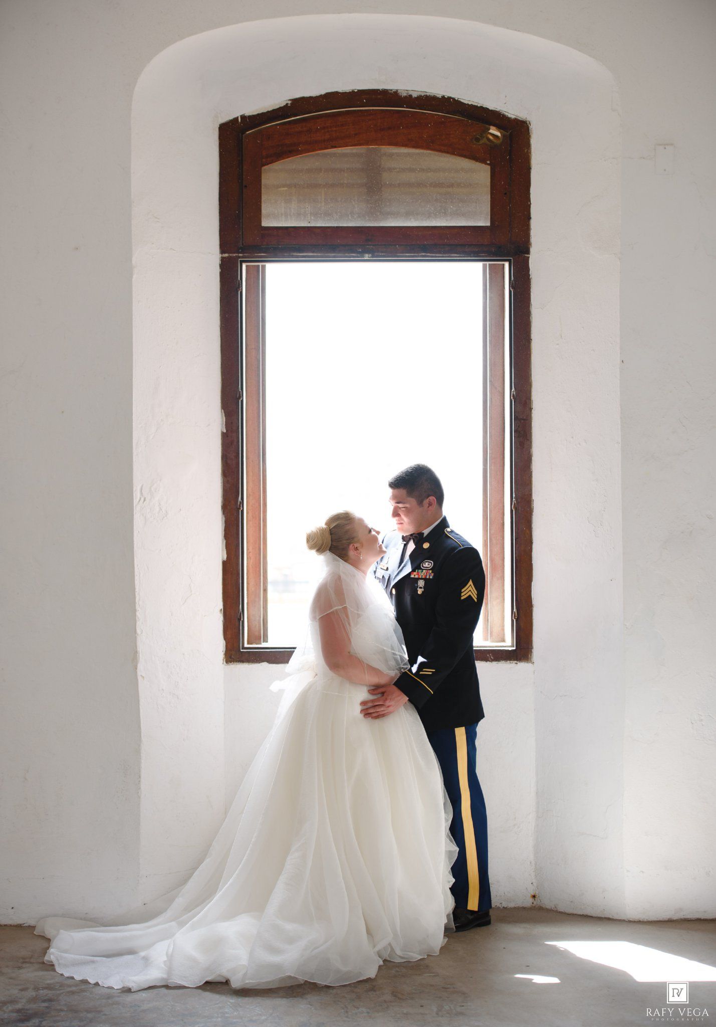 Military groom in dress uniform embraces bride in white ball gown before arched window in elegant wedding portrait.