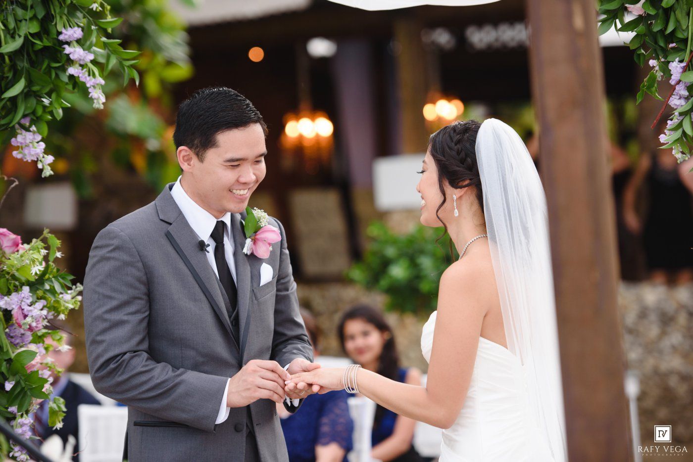 Groom places wedding ring on bride's finger during outdoor ceremony with floral arch backdrop.