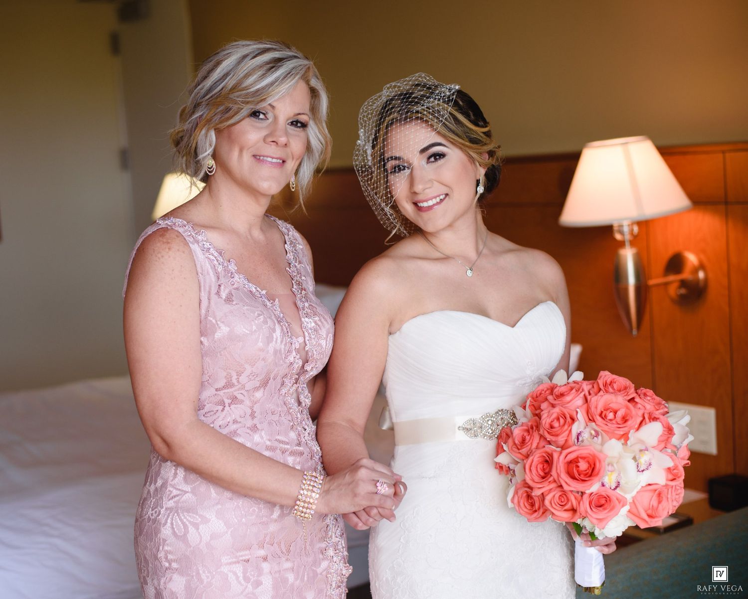 Bride in white strapless gown holding coral bouquet poses with woman in pink lace dress in hotel room.