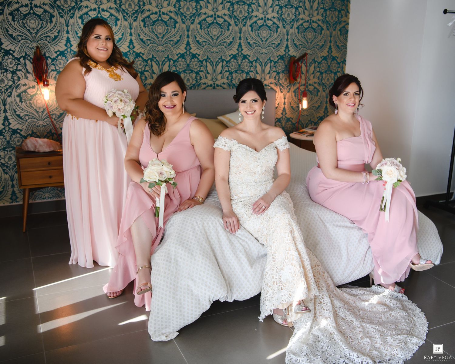 Bride in white lace gown poses with three bridesmaids in pink dresses holding bouquets in elegant room.