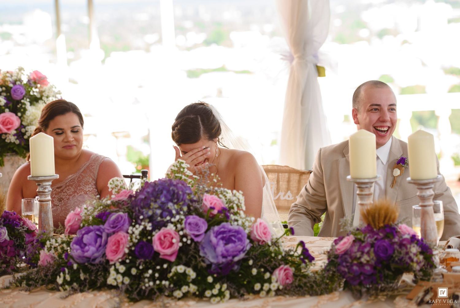 Bride and groom laughing joyfully at wedding reception table adorned with purple and pink floral arrangements.