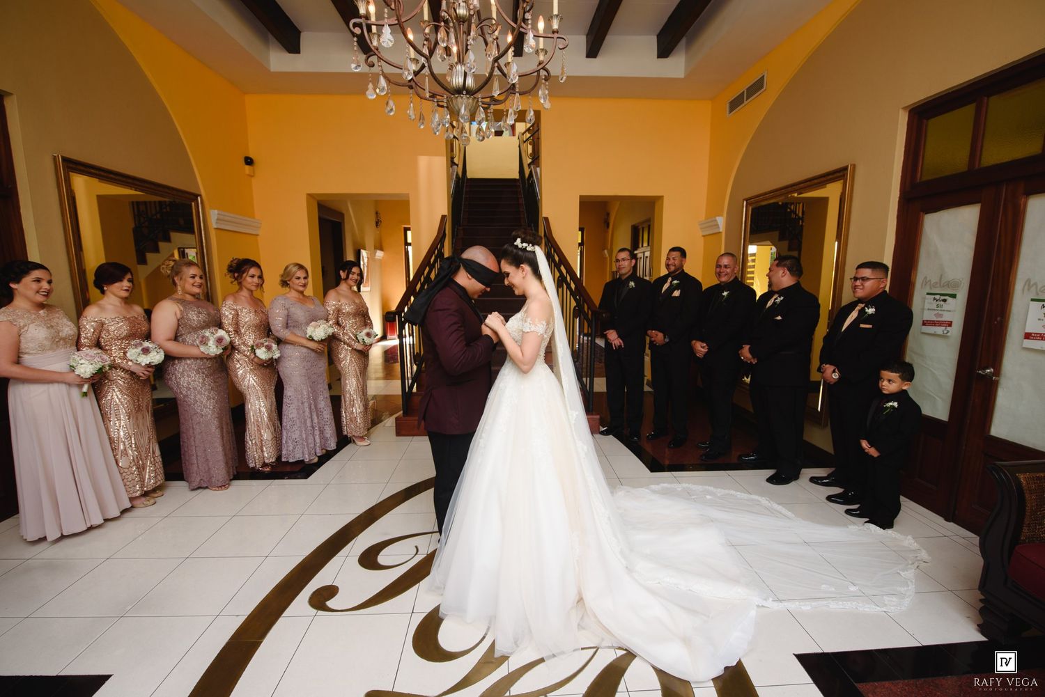 Bride and groom share first dance under chandelier with bridesmaids in blush gowns at wedding reception.