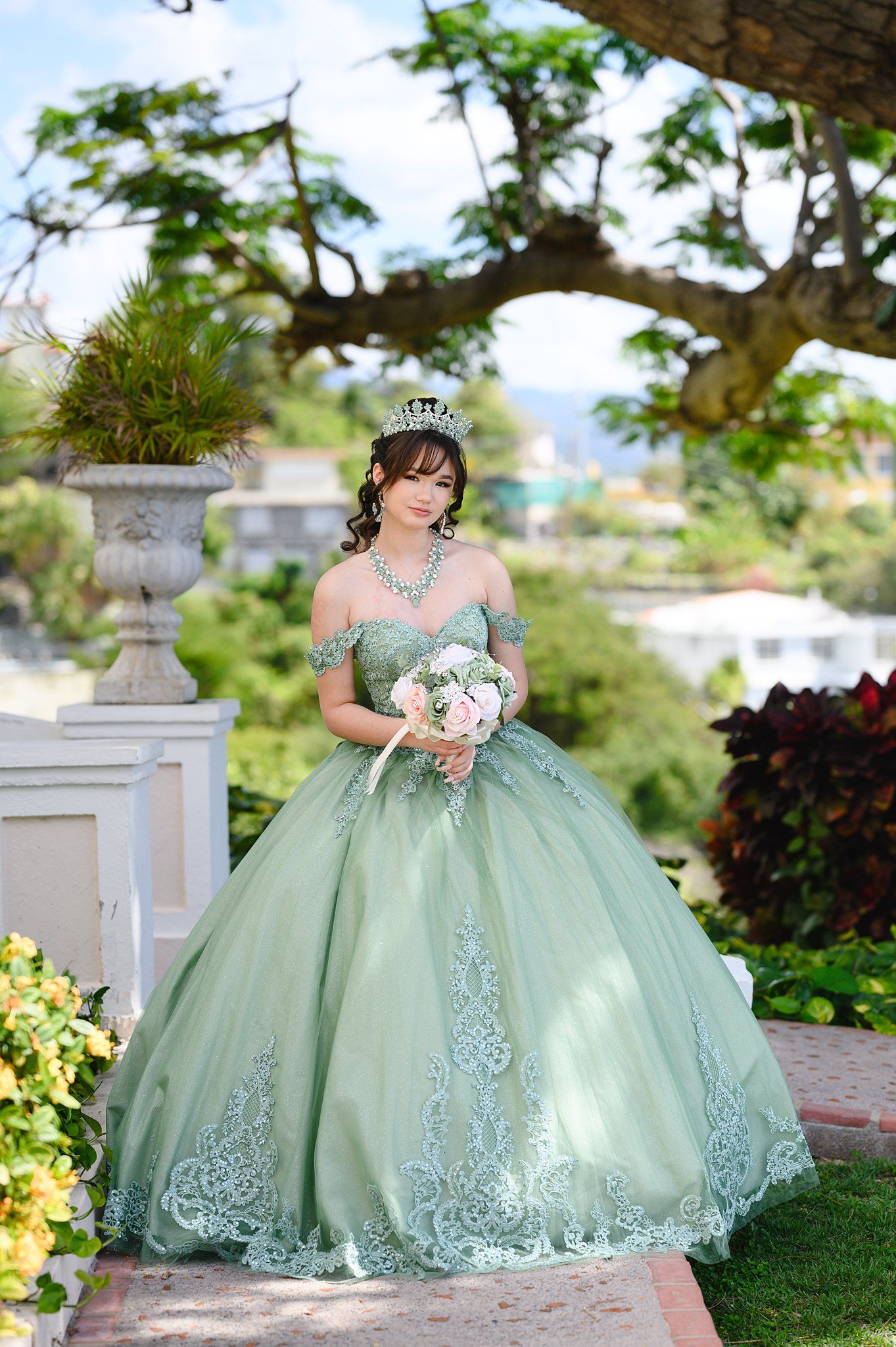 Young woman in sage green quinceañera ball gown with lace, crown, and bouquet poses in lush garden setting.