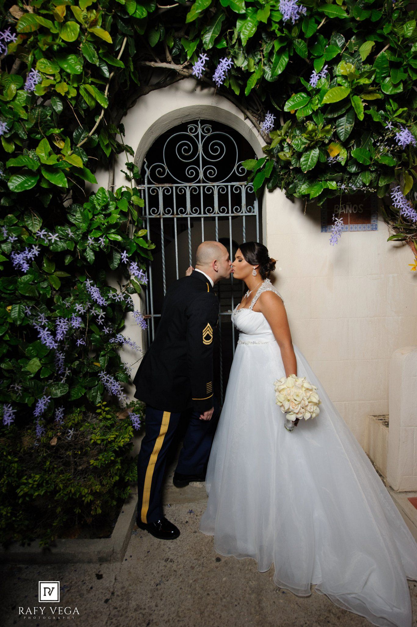 Military groom in dress uniform kisses bride in white ball gown beneath flowering vine archway at outdoor wedding.