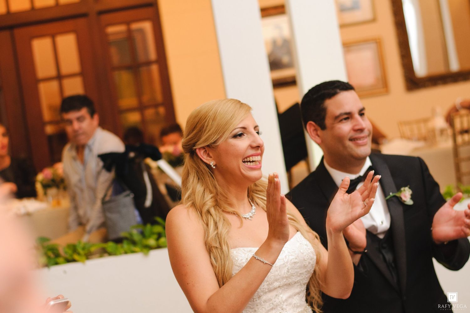 Bride and groom clapping and smiling joyfully at their wedding reception in an elegant venue.