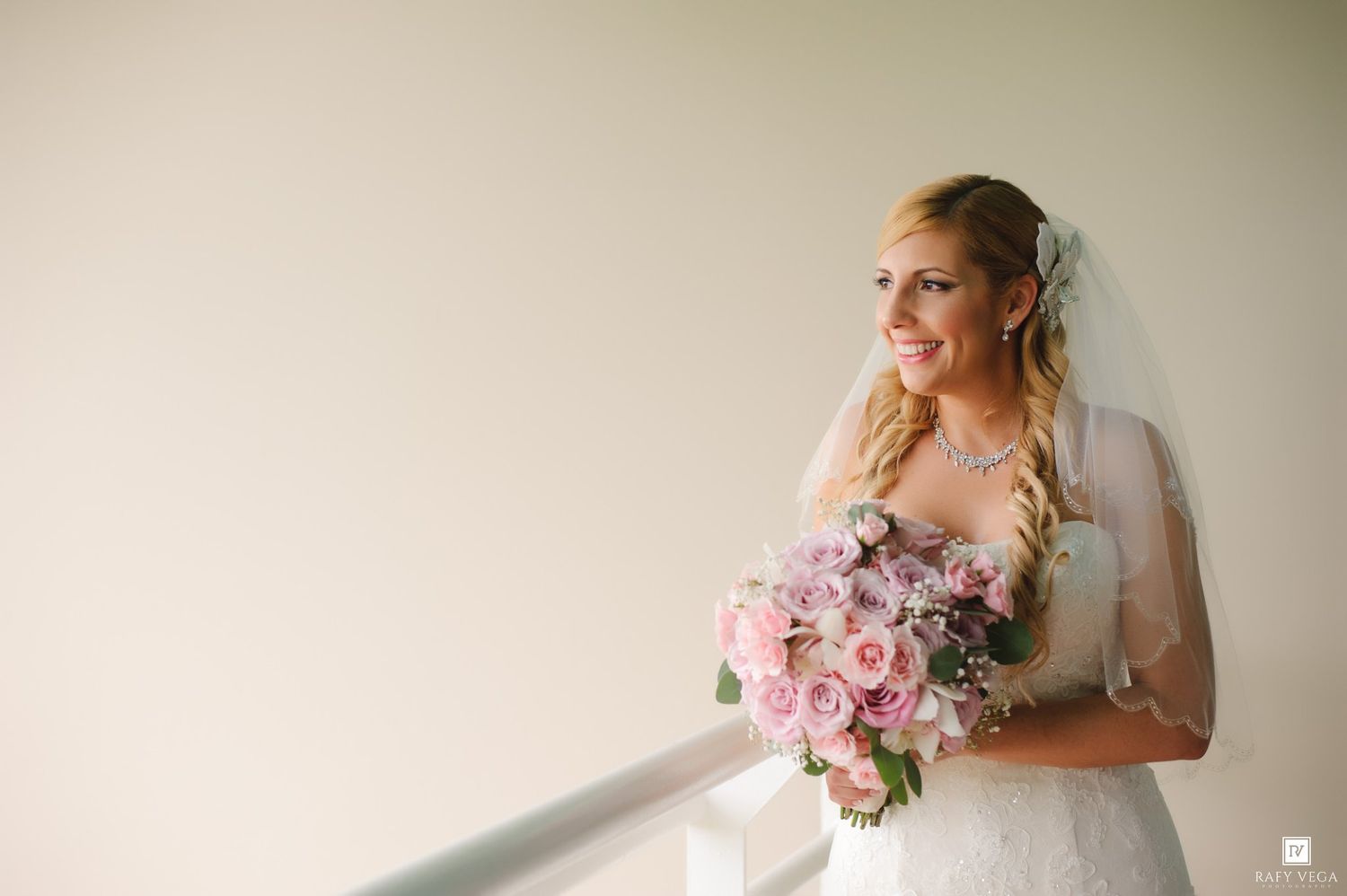 Smiling bride in white lace gown holding pink rose bouquet with veil, posing by window in soft natural light.