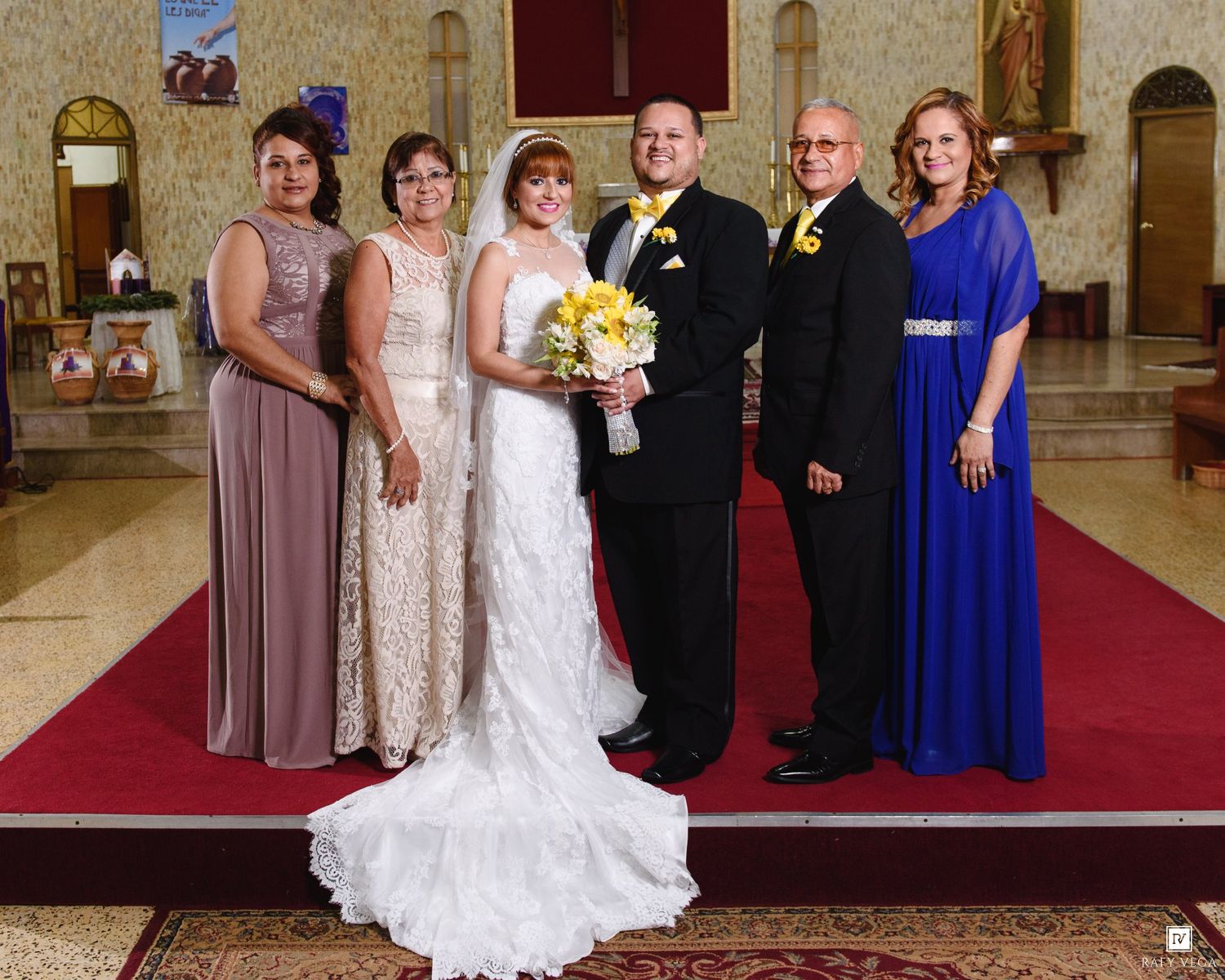 Wedding party poses in church, bride in white gown holding yellow bouquet, flanked by family in blue and gray attire.