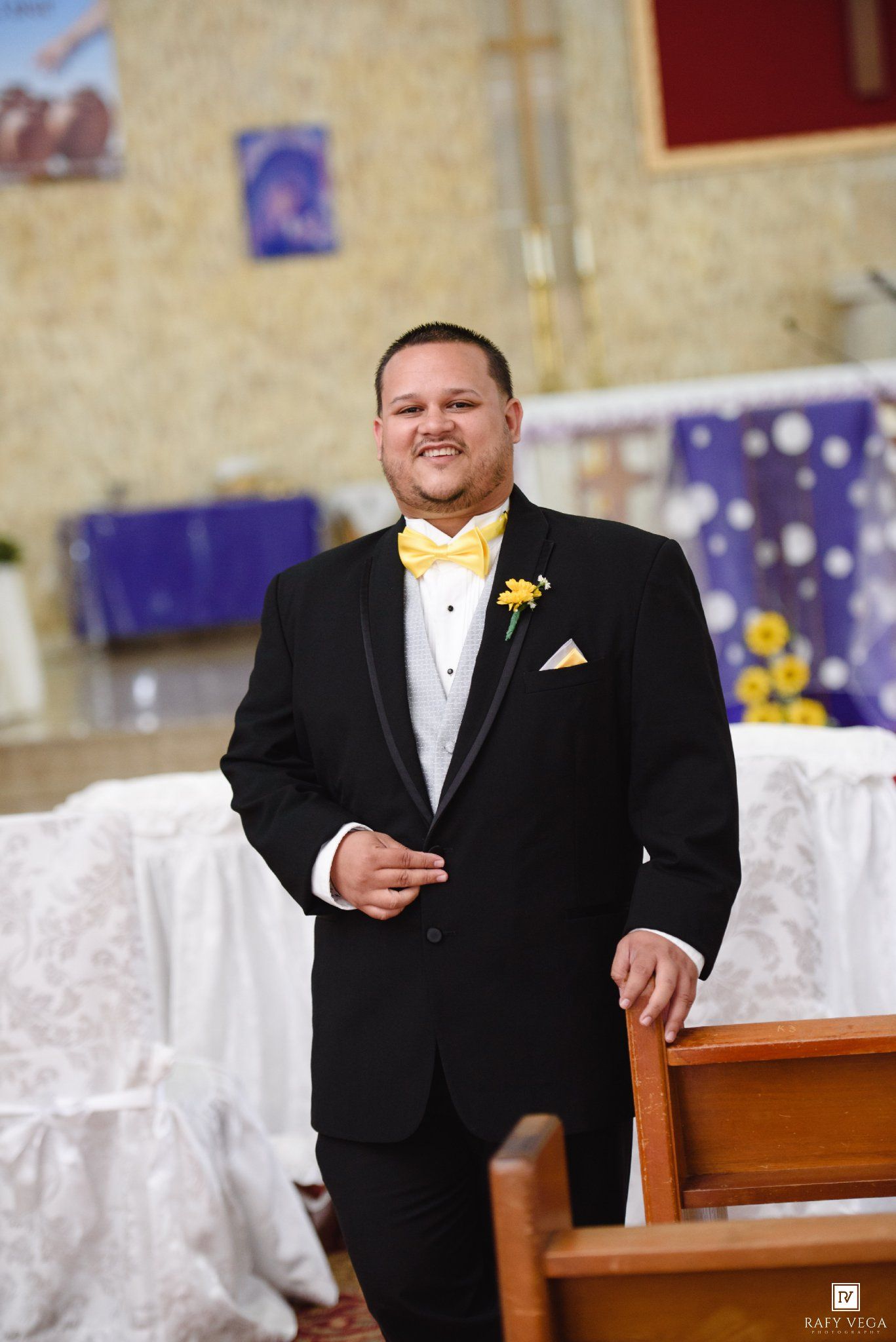 Smiling groom in black tuxedo with yellow bow tie and boutonniere standing in a decorated church.
